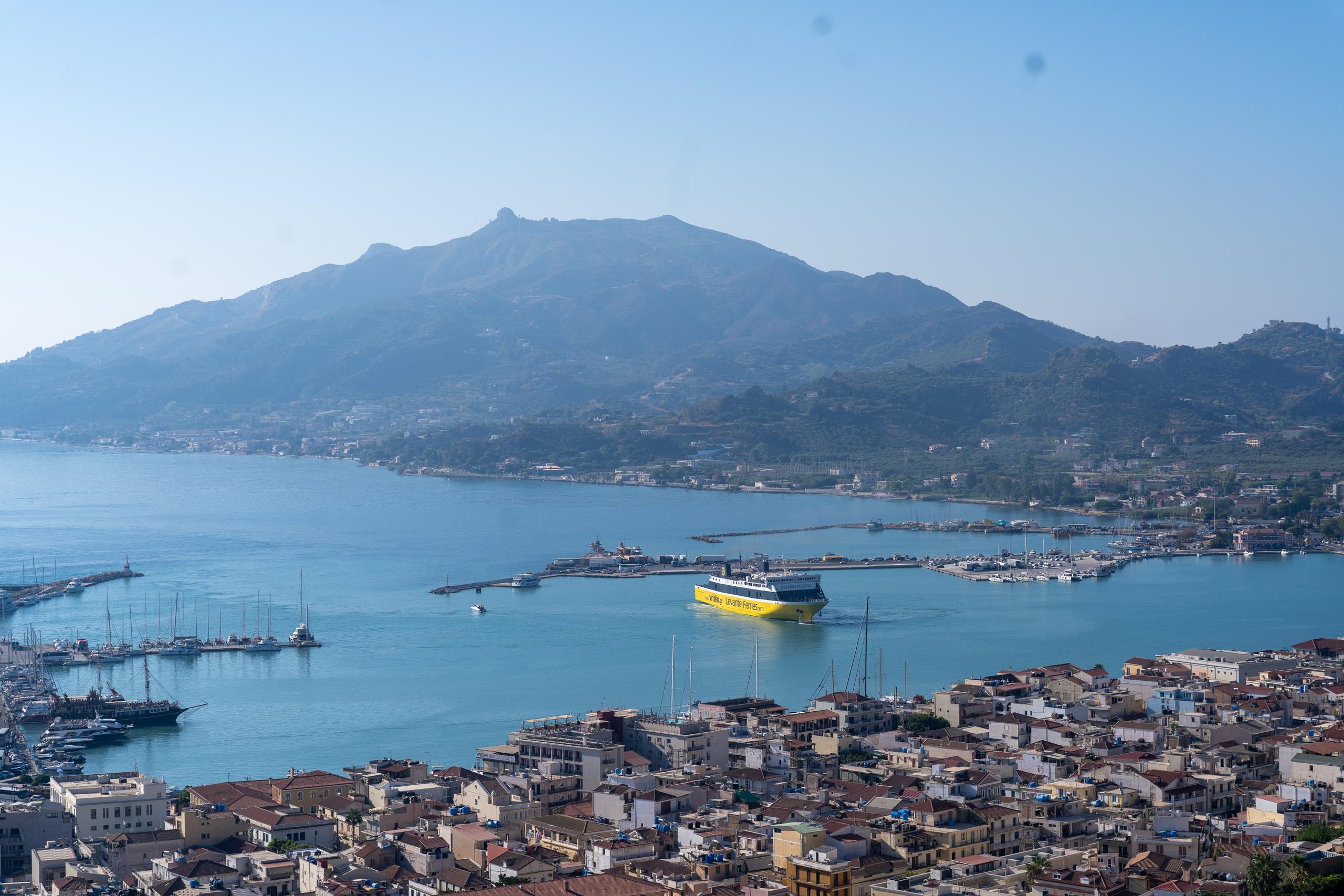 A high-angle view of the harbor city, with dense blocks of red-tiled roofs covering the bottom of the frame. A bright yellow ferry is sailing in the bay, and rows of sailboats and boats are parked in the harbor. Behind the city is a large mountain covered with vegetation and with rocky peaks on the top. The sky is light blue and misty.