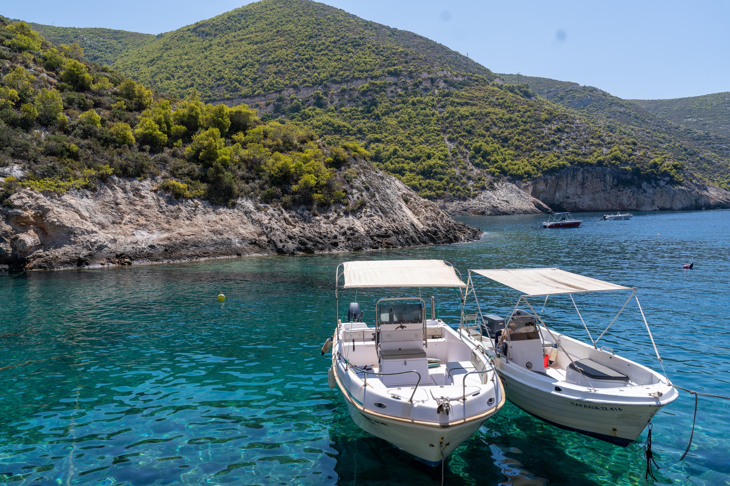 In the crystal clear turquoise bay, light-colored rocks and light spots can be seen under the water; two white boats with beige awnings are parked side by side in the front of the frame, and in the background are green hillsides covered with shrubs and exposed light-colored rock walls. There are two boats in the distance at the rear right.