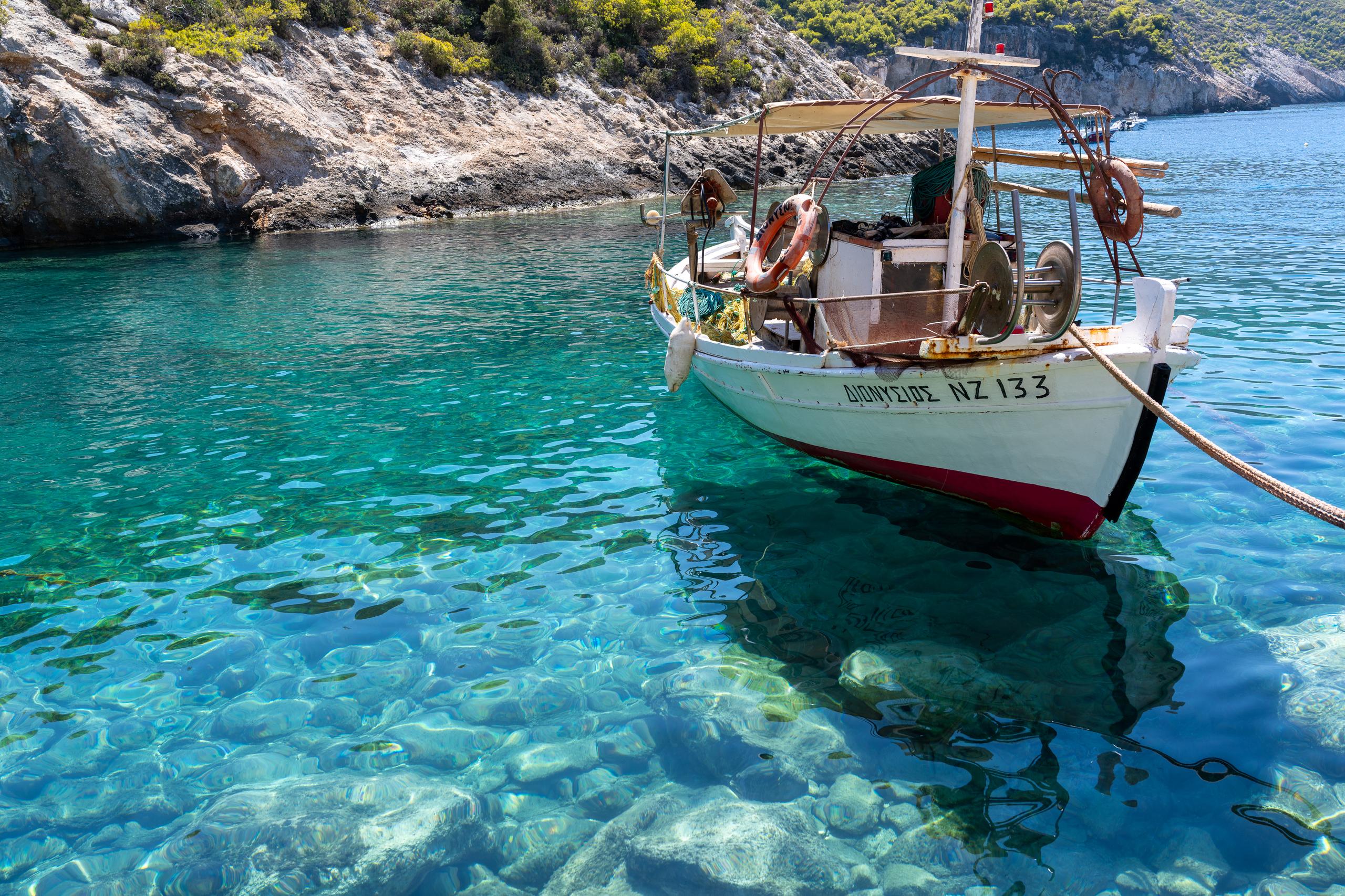 A traditional wooden boat with a white hull and a red waterline is parked on a rope in the clear turquoise shallow water. A simple awning is erected on the boat, and orange lifebuoys and fishing gear are hung on the boat. Through the water, you can clearly see the underwater rocks and the reflection of the boat's hull. The background is light-colored rocky shores and shrub hillsides.
