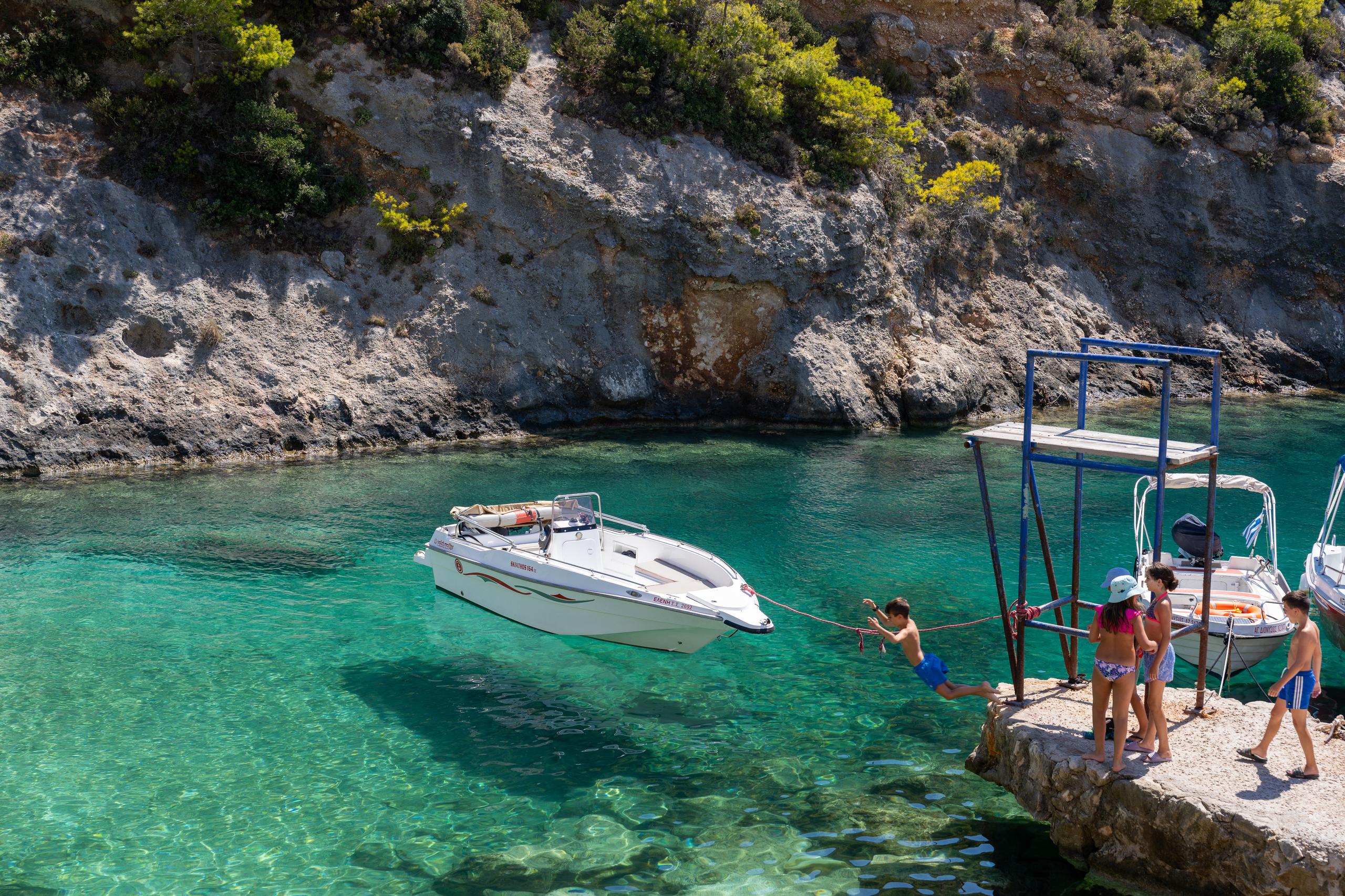 A rocky shore bay in the sun: There is a blue metal support on a small stone pier on the right side of the frame. Four children in swimsuits are standing on the edge of the pier. One of them is hanging on the water, holding on to a rope, and the other is walking down the stone steps; on the left is a white speedboat parked on the water. The water is turquoise and transparent, and the background is light-colored rock walls and shrubs full of caves.