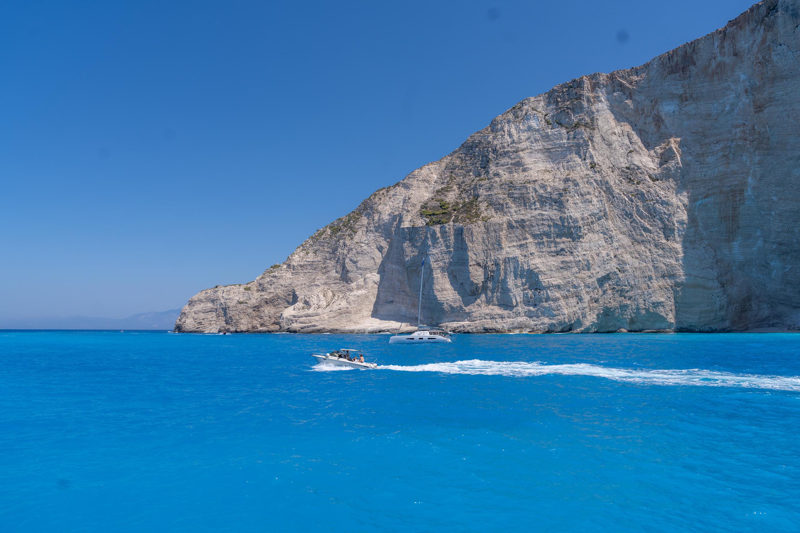 Under the deep blue sky, an almost vertical white limestone cliff extends from the right side of the frame, with clear rock texture. On the blue sea below the cliff, a speedboat drags a long white trail to the right, and there is a sailboat next to it. On the distant horizon is a faint outline of land, and the water is a saturated deep blue.