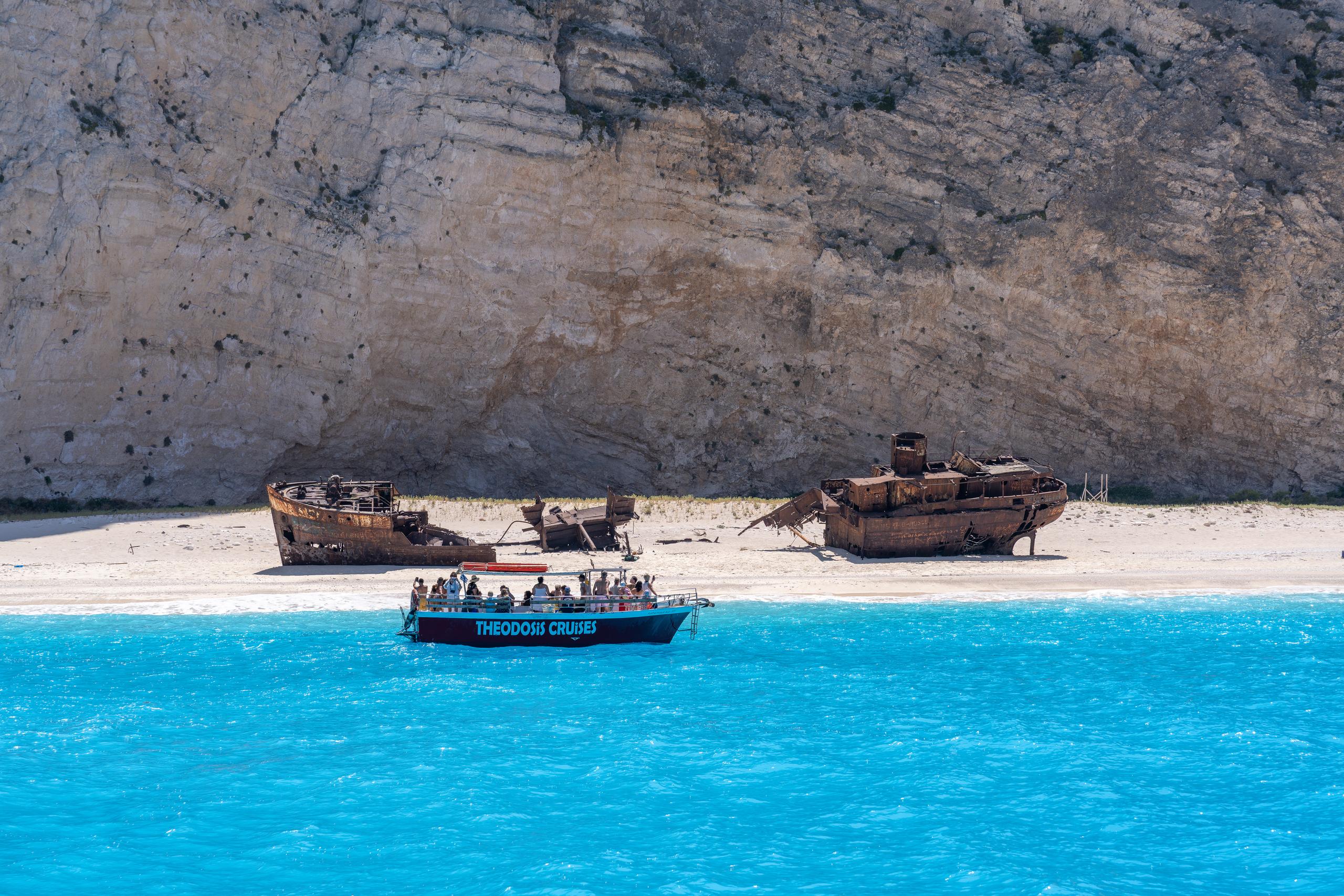 A large rusty sunken ship was broken into several pieces and stranded on the white pebble beach. The rusty red hull formed a strong contrast with the white beach surface; in the foreground was a blue boat parked on the blue sea with white writing on the hull and the deck was full of tourists; behind it was a whole light-colored rock wall with distinct layers.