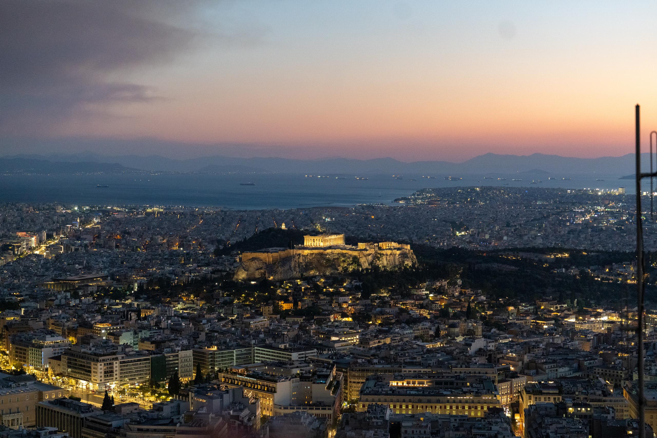 Panoramic view of the city at twilight: Dense city lights fill the lower half of the frame, with the ruins of an ancient temple illuminated on top of a rocky hill in the center; in the distance are the dark blue bay, port lights and continuous mountain shadows, with an orange-pink glow band across the horizon, and the rest of the sky is gray-purple.