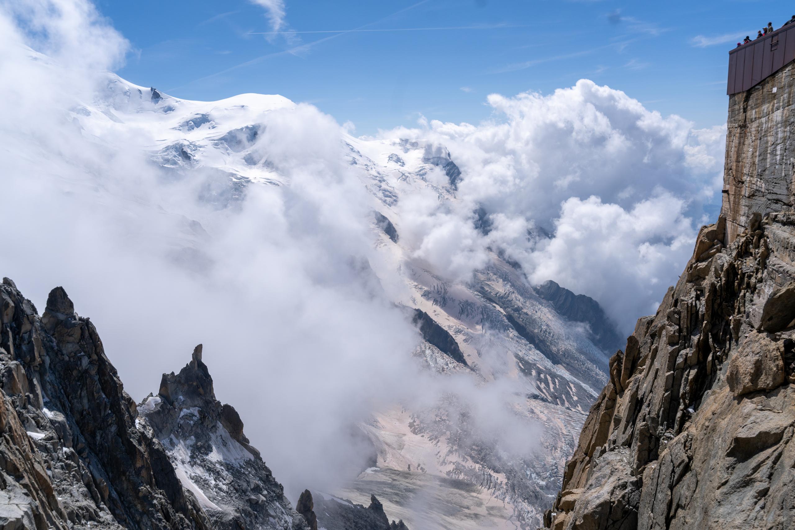 Clouds and mist rise from the valley, and in the distance are the peaks and blue sky covered with thick snow and glaciers. The lower left and right sides of the frame are dark and sharp rock ridges; the upper right corner of the viewing platform built on the rock wall is exposed, with several people standing on the platform.