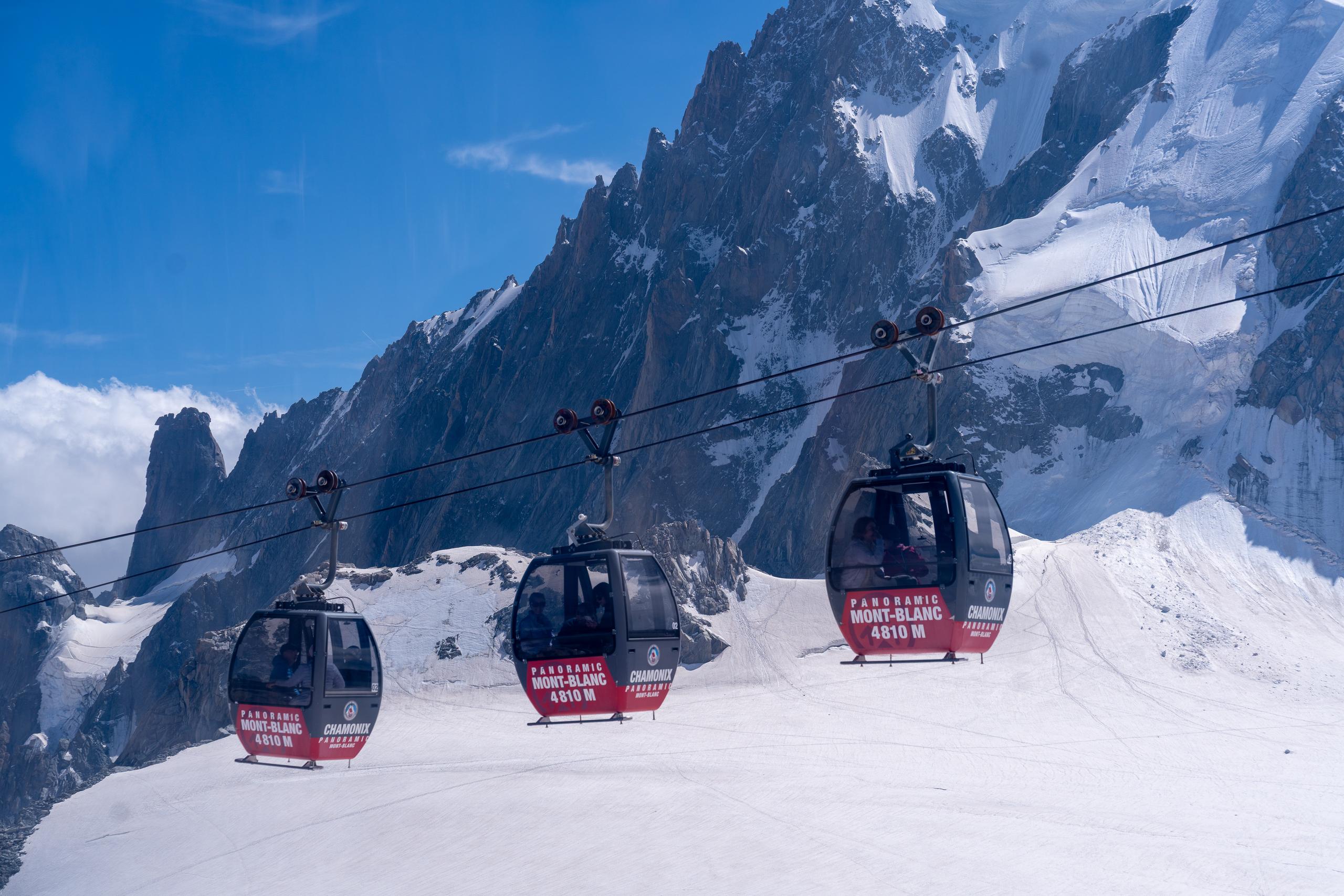 Three cable cars with glass viewing cabins are suspended on the cableway above the snow slope, arranged from left to right. The lower part of the car is red and printed with white words; behind it are snow-covered rock walls and glacier fissures, and the sky is dark blue.