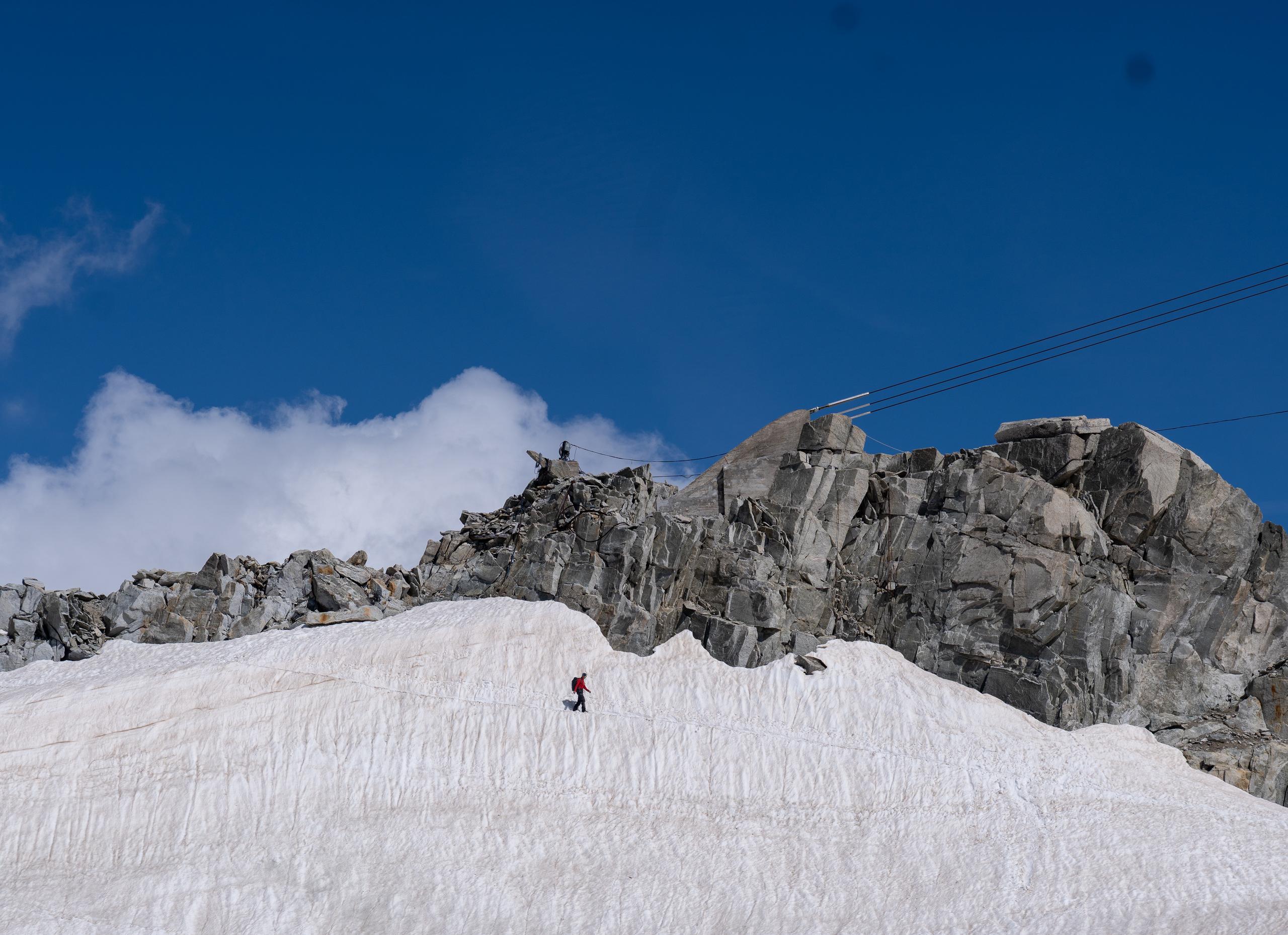 Under the dark blue sky, a snowfield full of folds and ravines extends from the bottom of the frame. In the center of the snowfield is a man wearing a red coat and carrying a backpack, walking alone on the snow; above the snowfield is a ridge of dark gray giant rocks, and several cables slant across the sky on the upper right.