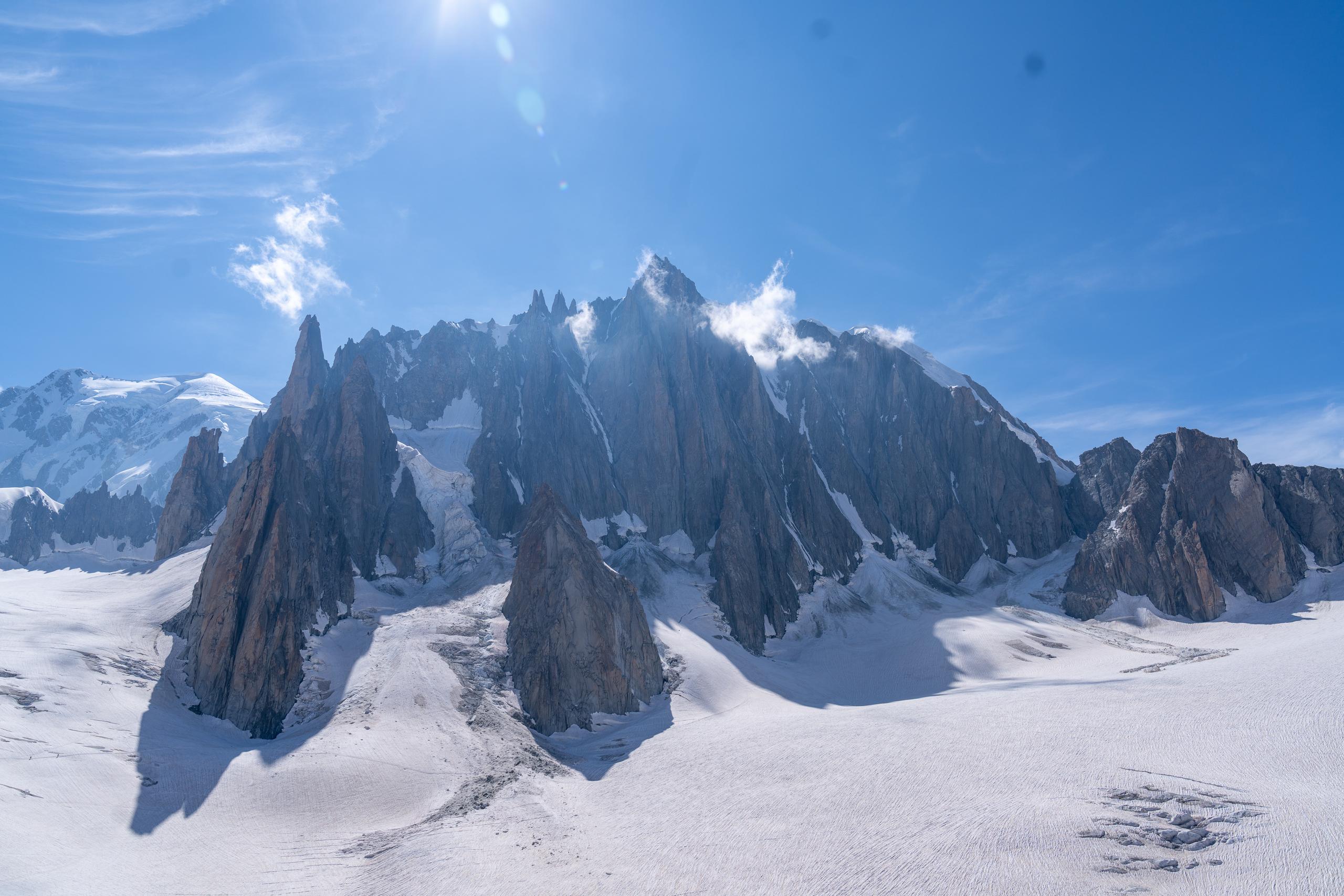 On the vast glacial snowfield, a row of dark cone-shaped rock peaks stand like sawtooths, with some rock walls covered with snow and ice. There is another snow-capped mountain in the distance on the left. There are strong sun spots and thin clouds at the top of the frame, and the long shadows of the rock peaks are trailed on the snow.