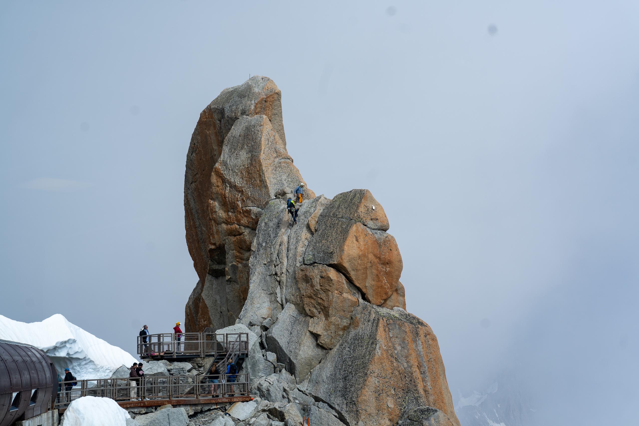 Against the foggy gray background, a huge granite pillar stands. The rock surface is yellow-brown and off-white. Two climbers wearing colorful clothes are climbing up on the lower right side of the rock pillar. Several tourists are standing on the metal viewing platform on the lower left side. Below the platform is the top of a cylindrical building.