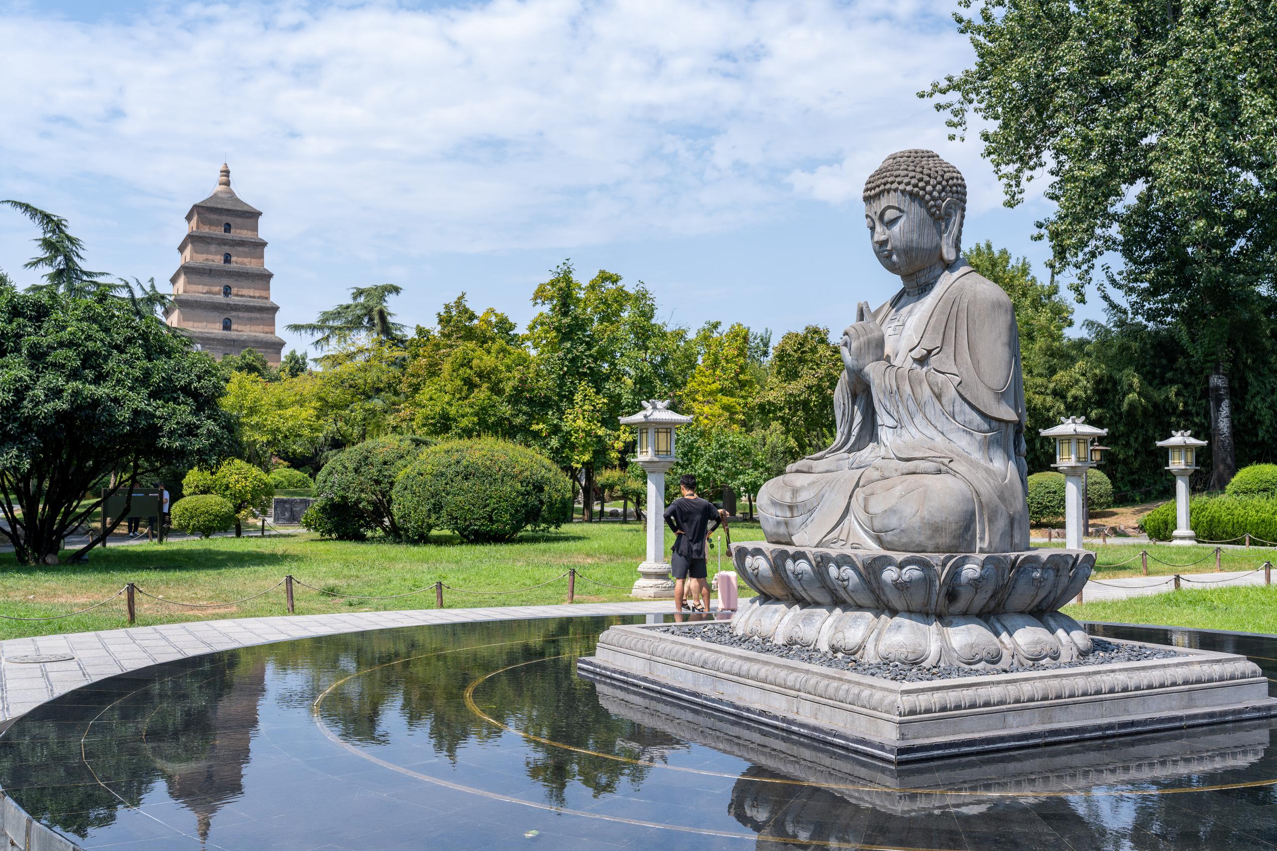A gray stone sculpture of Buddha sitting in lotus position on a lotus seat is located in front of the right side of the frame. The Buddha statue is perched on a square stone platform. In front of the frame is a shallow round pool, with the water reflecting the sky and trees. The distant view on the left is the square brick tower. The middle ground is a neatly trimmed lawn and bushes. There are several white stone lamps on the grass. A man in black stands beside the Buddha statue and watches.