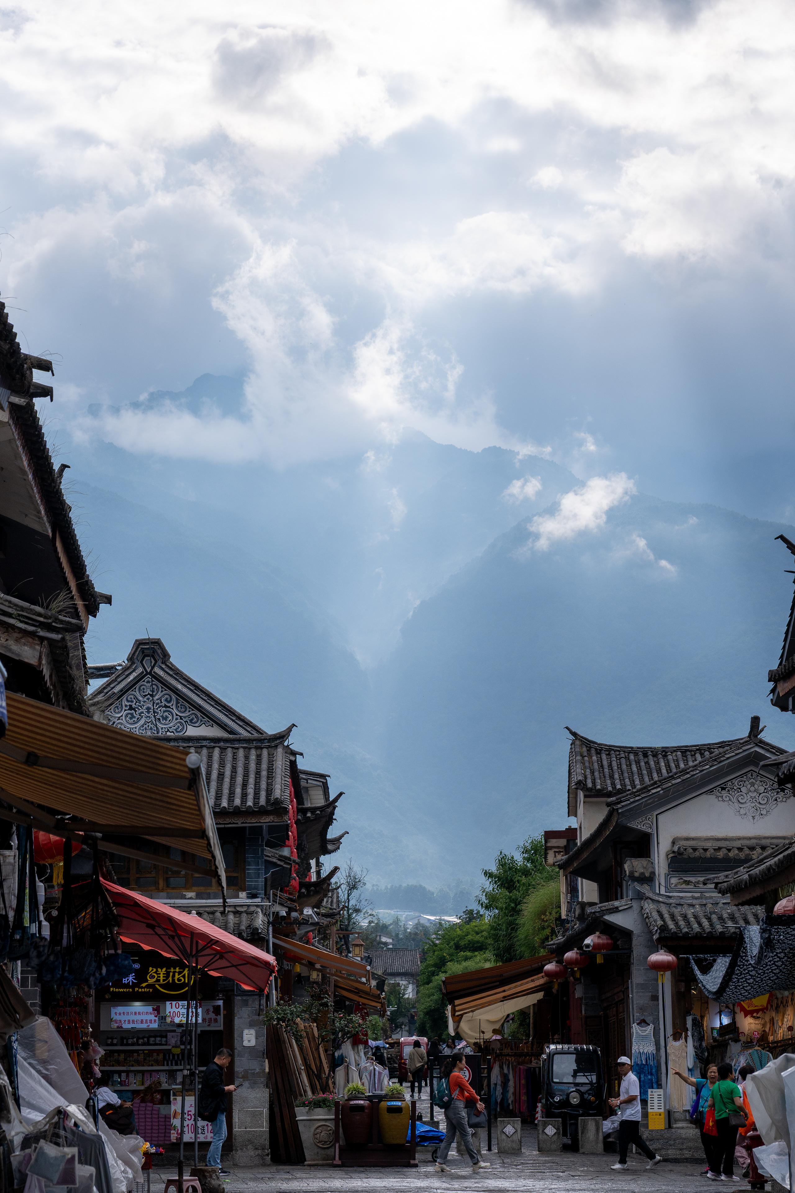 The street scene of an ancient city commercial street, with gray tiles, angular eaves, and traditional shops with wooden doors and windows on both sides. There are awnings and red parasols in front of the shops. Red lanterns are hung on the street, and goods are piled up. Pedestrians are walking in the alley. The end of the street faces a huge mountain shrouded in clouds and mist. White clouds float on the mountainside, and the sky is filled with thick clouds.