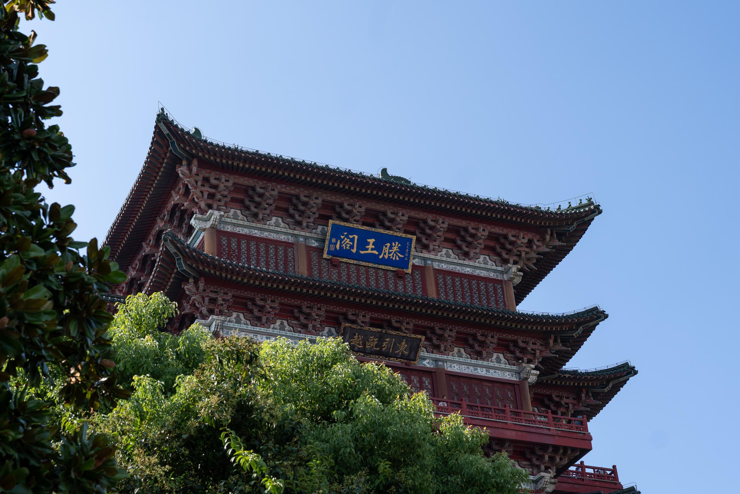 Looking up at a multi-story Chinese-style pavilion, dark red pillars and beams, multi-layered gray tile eaves are stacked up one after another. There are complicated brackets and painted decorations under the eaves, and there are ridge animals on the ridge. There is a blue-based gold plaque hanging on the front of the pavilion, and there is a dark plaque below; the foreground and left side are covered by dense green leaves, and the sky is clean and light blue.