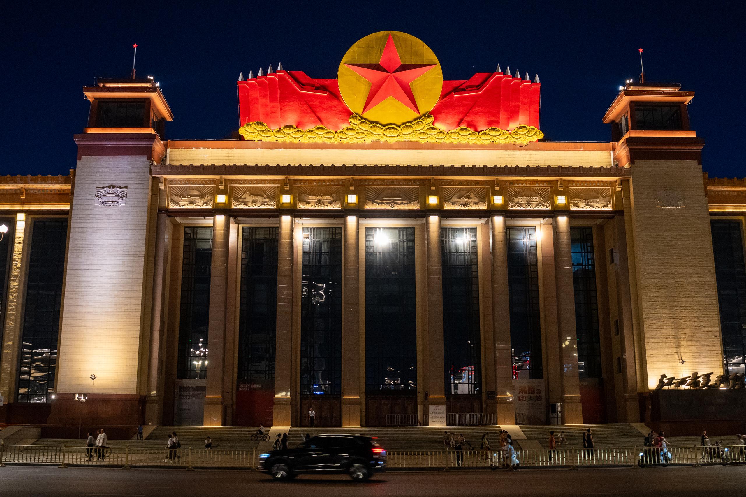 The front facade of a large public building under the night view. Square columns are arranged on the tall beige wall, and the glass windows between the columns reveal warm light. There is a red and gold circular decoration and a red flag-shaped relief in the center of the top of the building. There is a turret with a square tower on each side. There are people walking on the steps in front of the building, and there are vehicles passing by on the road below.