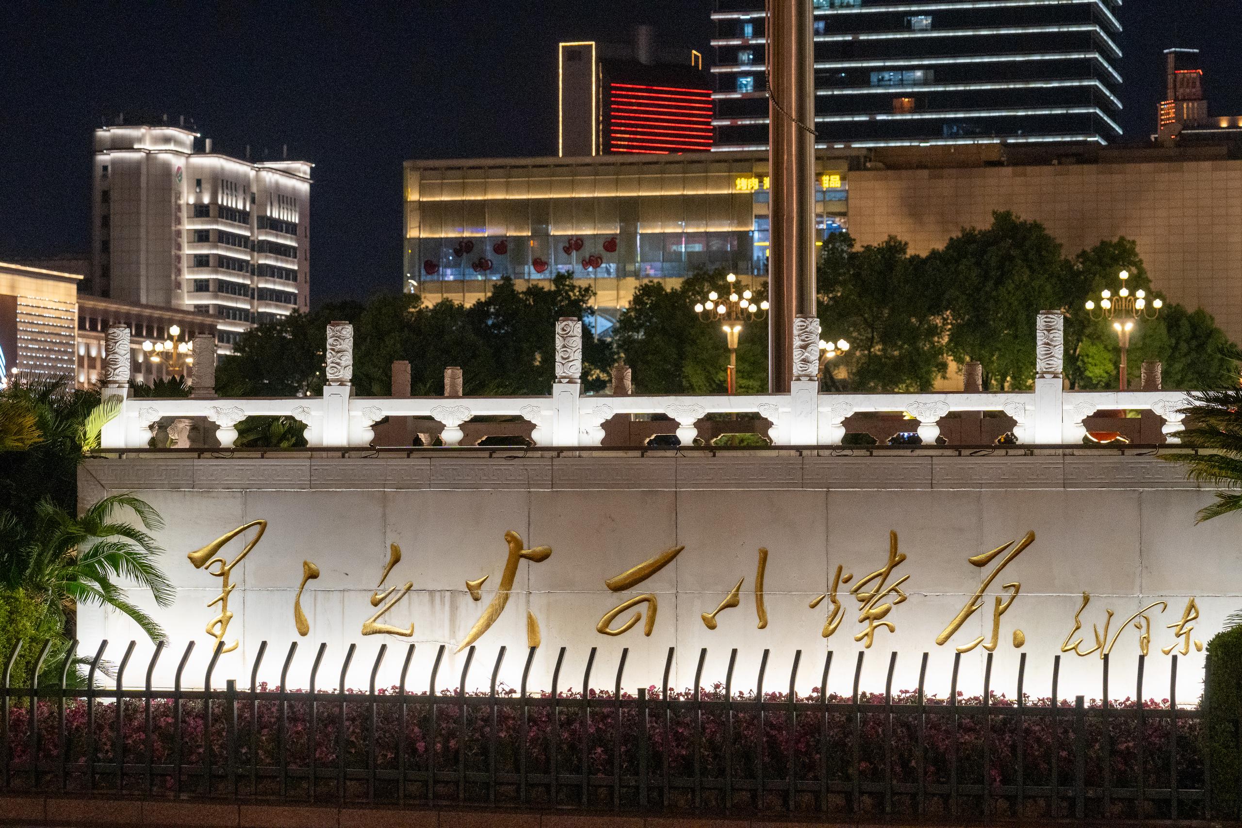 In the corner of the square at night, a white stone guardrail and a low wall run across the middle of the frame. The low wall is inlaid with large golden inscriptions. In front of the wall are flower beds with railings and rows of shrubs. Behind the low wall is a city night scene, with many modern high-rise buildings and illuminated shops visible. One of the floors has a red horizontal light strip and several spherical street lights.