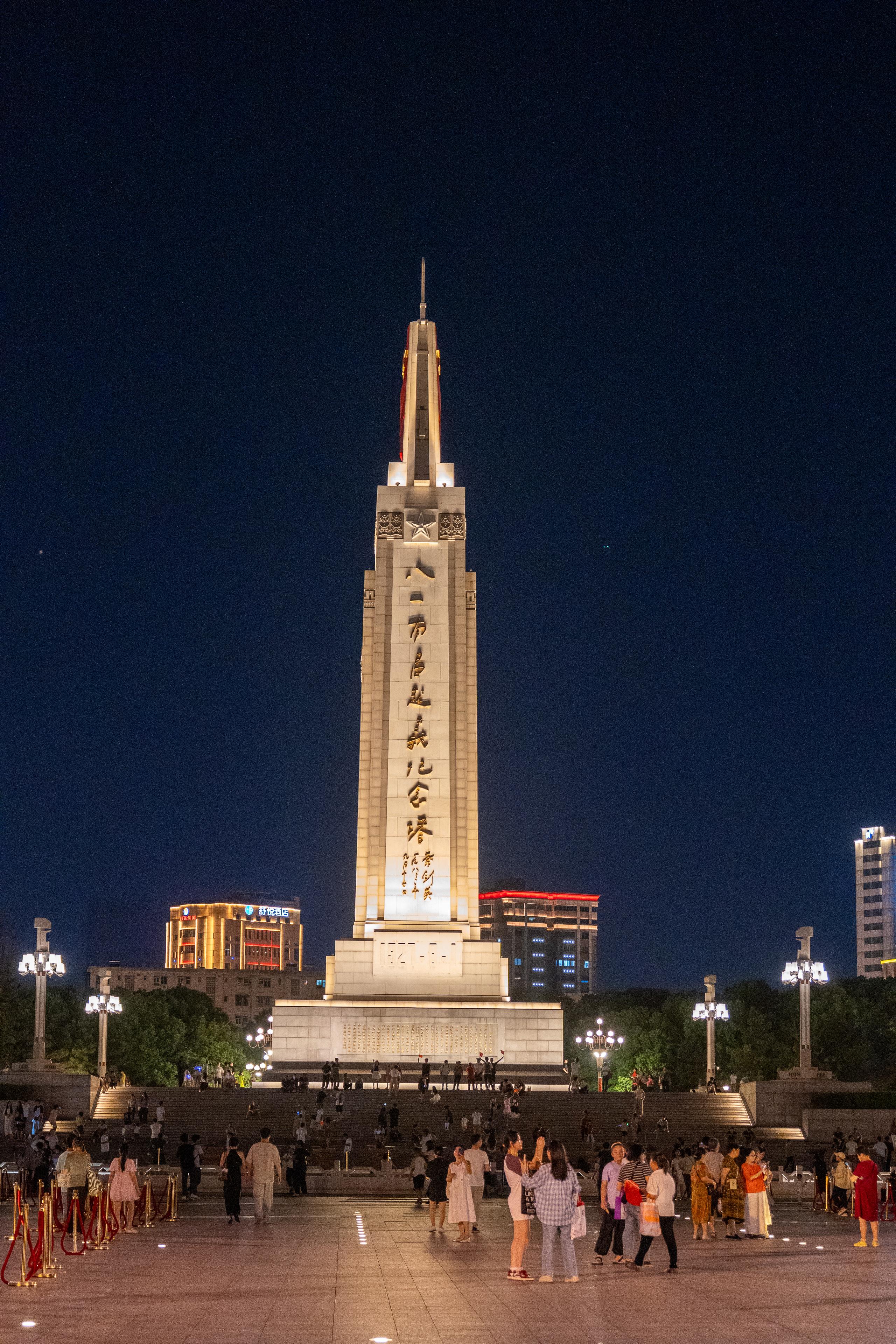 In the memorial square at night, in the center of the frame is a towering obelisk-style memorial tower. The tower is engraved with large golden characters in vertical rows, and the top is decorated with a spire and red decoration. The base of the tower is a multi-layered stone step. Many people walking and taking photos gather in the square in front of the tower. Street lamps with spherical lampshades stand on both sides. High-rise buildings with bright lights can be seen in the distance, and the sky is dark blue.