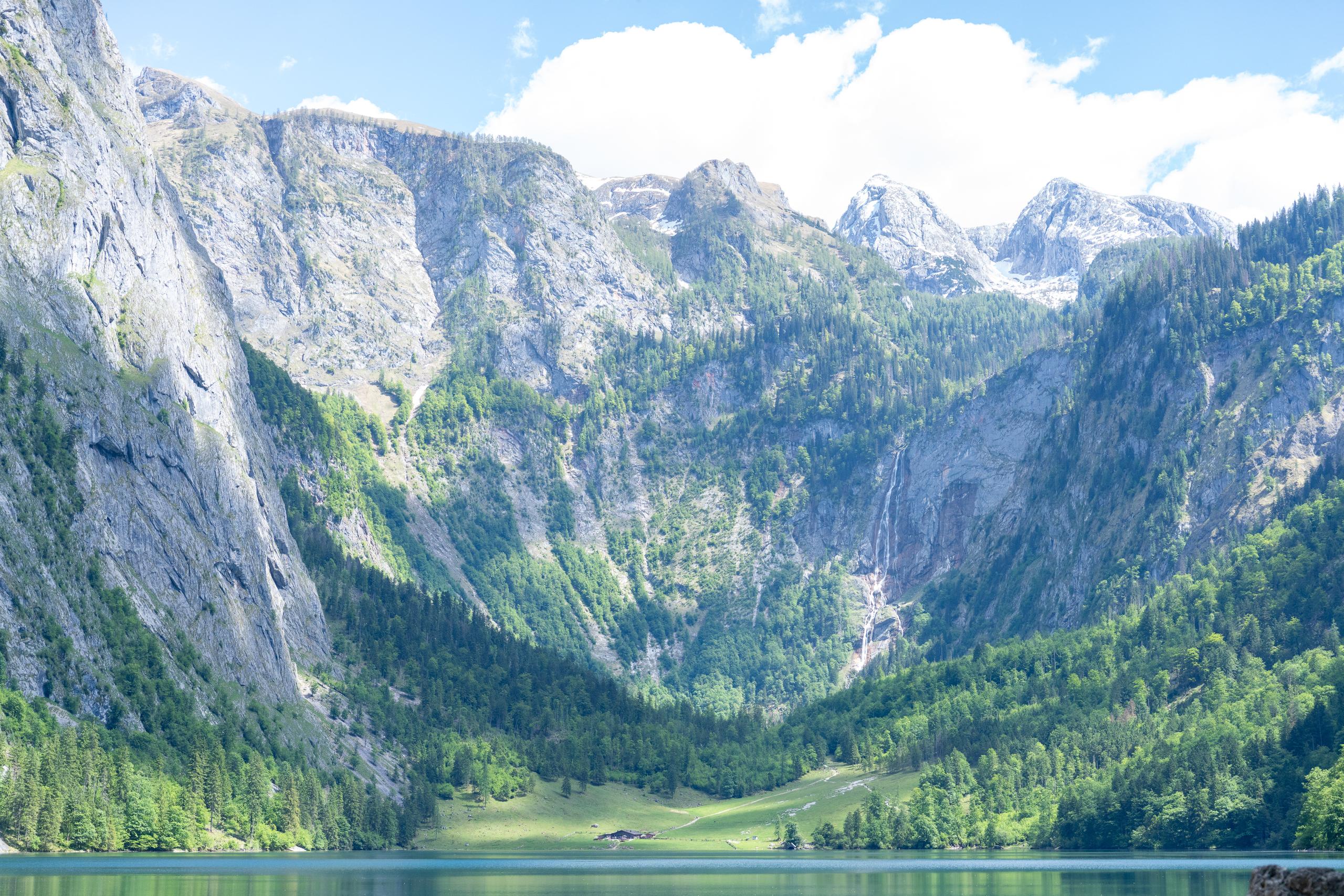 A broad panoramic view of an alpine lake: the dark blue surface of the lake runs across the lower part of the frame. On the left side of the lake shore is a steep, almost vertical light gray rock wall, and on the right side of the mountain there is a slender waterfall pouring down from a high place; the hillside is covered with dark green coniferous forest and grassland, and there is a small wooden house on the lakeside grassland; the top of the mountain is covered by white clouds, and the sky is filled with cumulus clouds.