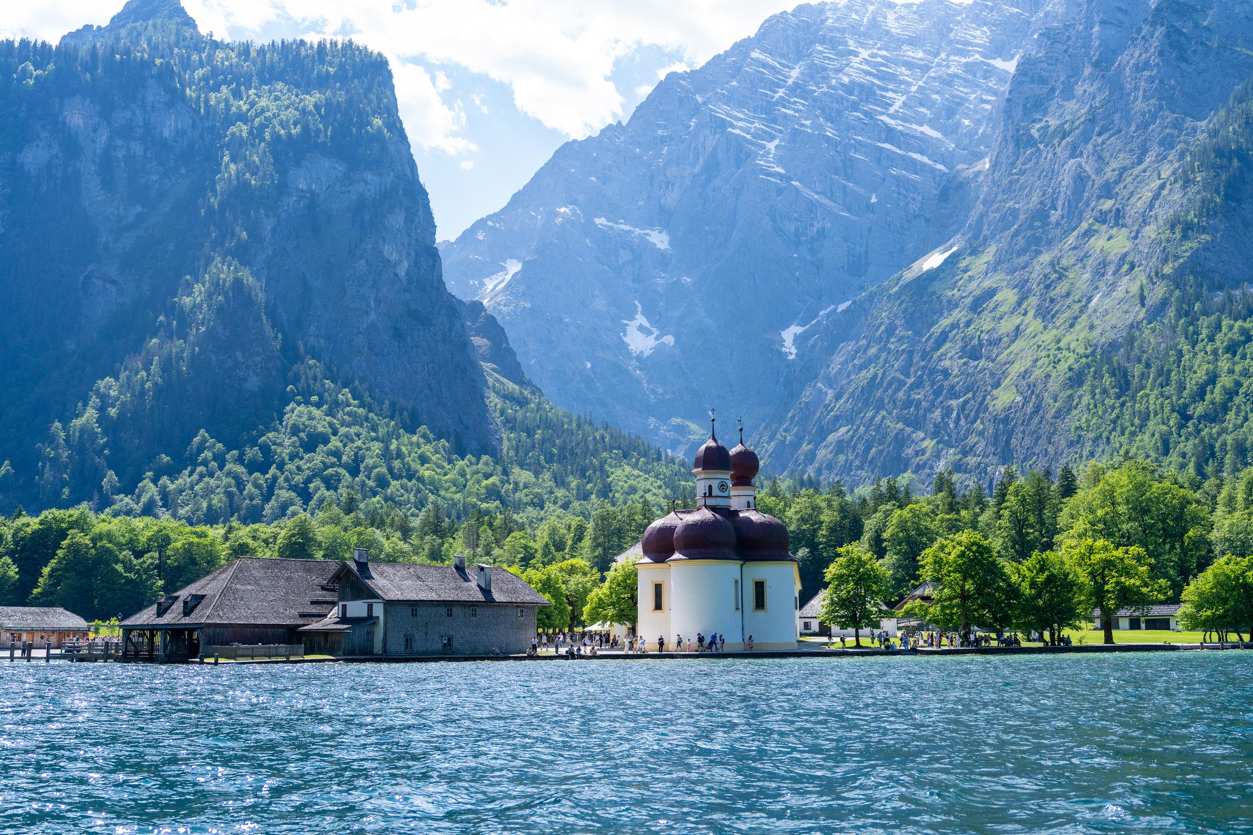 There is a small church by the lake with white walls and a dark red onion-shaped dome, surrounded by green trees. On the left side of the church is a rectangular building with a dark pitched roof and a wooden corridor. Many tourists gather on the shore. The lake is blue and rippled. In the background are two towering rock peaks, with white snow remaining on the mountains and clouds and mist in the valleys.