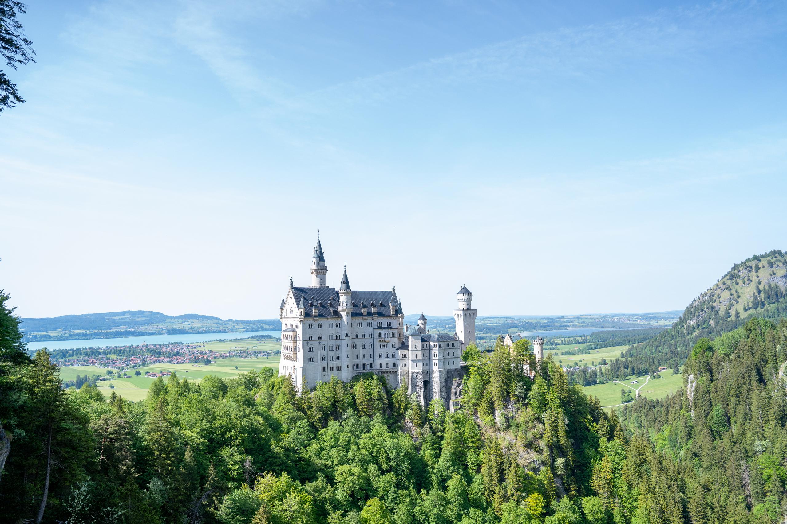 A white castle-like building complex is located on a ridge covered with dark green forest, with multiple spiers and a tall square tower, with light-colored exterior walls and dark roofs; behind the castle are open plains, villages and towns, and a light blue lake. On the right is a green hill covered with trees, and the sky is light blue with thin clouds.