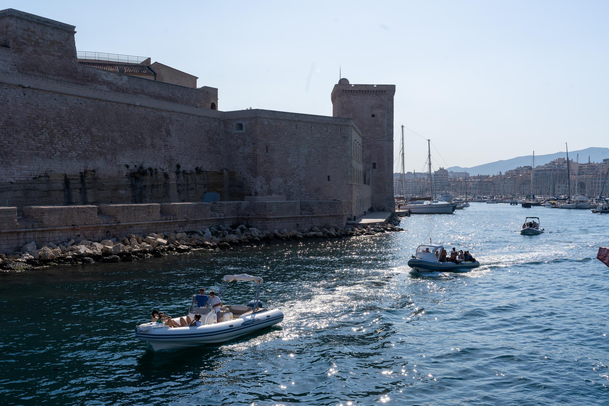The ancient stone walls and square towers on one side of the harbor are mottled and have a piled stone revetment at the bottom. Sunlight shines obliquely on the stone walls. Three rubber boats are carrying people in the harbor. The sea surface shines with dense silver reflections in the sunlight. In the distance are rows of moored sailboats and the city skyline.