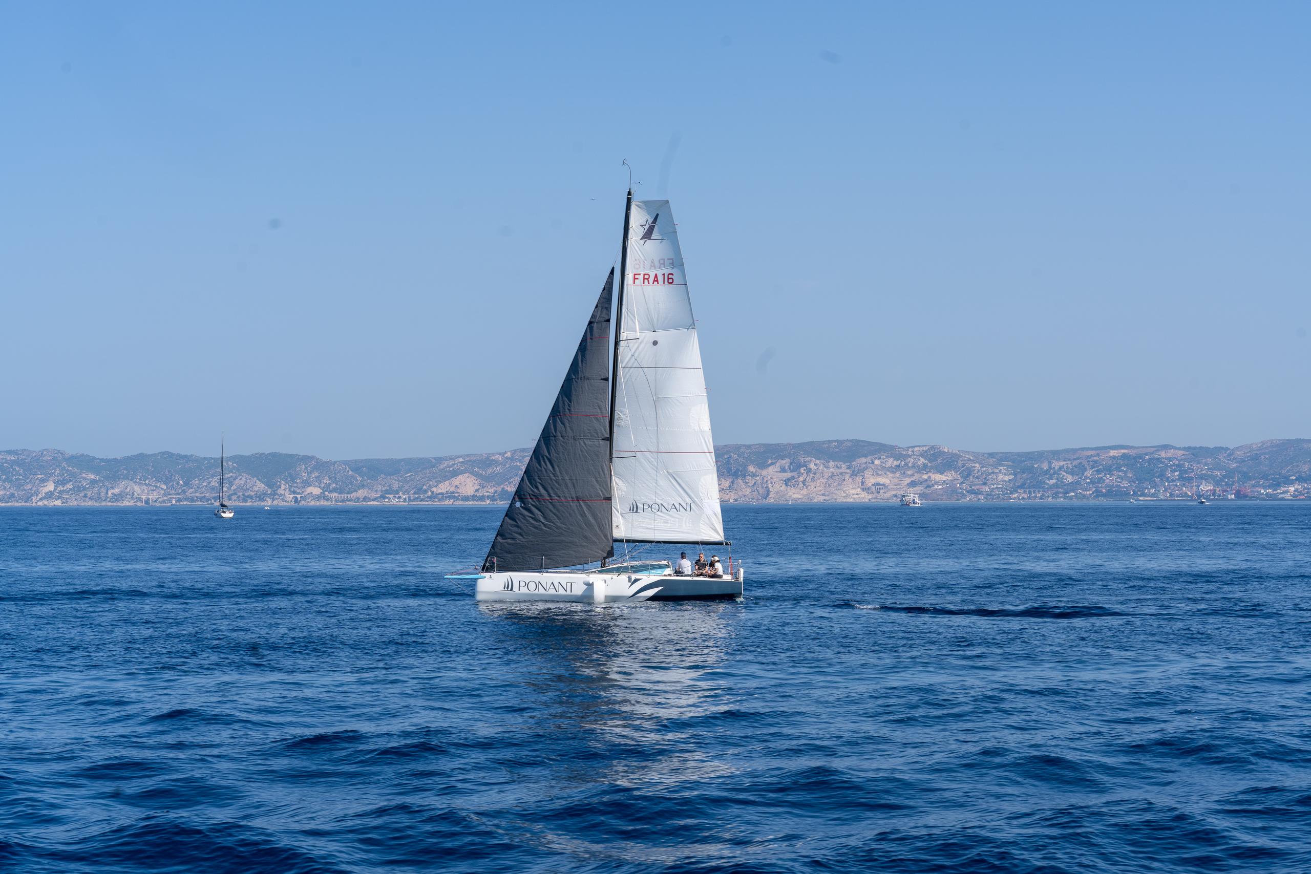 A white catamaran is sailing on the clear sea. The sails are half dark gray and half white. There are letter numbers and patterns on the sails. There are several people sitting on the deck. In the distance are the undulating light brown coastal hills, and the water is dark blue with fine ripples.