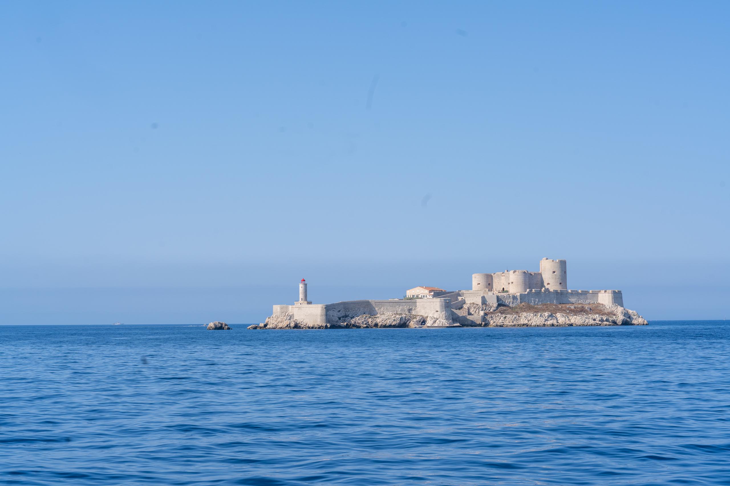 On the open sea, a small rocky island stands alone. On the island is a white stone fortress with multiple cylindrical towers and a wall. There is a small lighthouse with a red top on the left end of the island. It is surrounded by only dark blue sea and clean light blue sky. In the distance, there is a layer of mist floating at the junction of sea and sky.