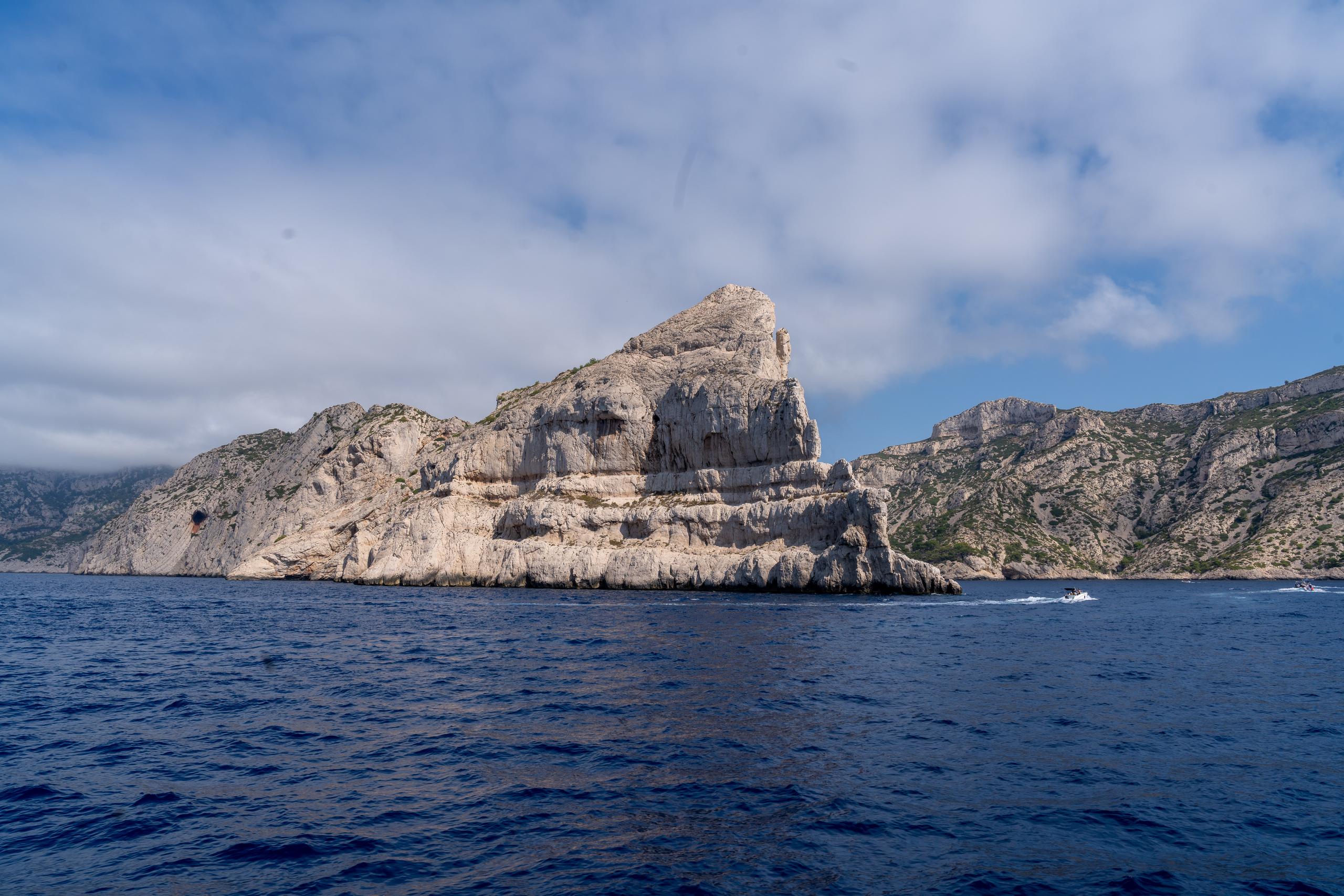 The white limestone cliffs on the sea surface are layered, with horizontal grooves and cracks formed by weathering on the surface. They extend from the left to the promontory on the right. There are scattered green shrubs growing on the ridges. Below the cliffs is the dark blue sea. In the lower right corner of the frame, there are white wakes left by two speedboats passing by, and the sky is filled with white clouds.