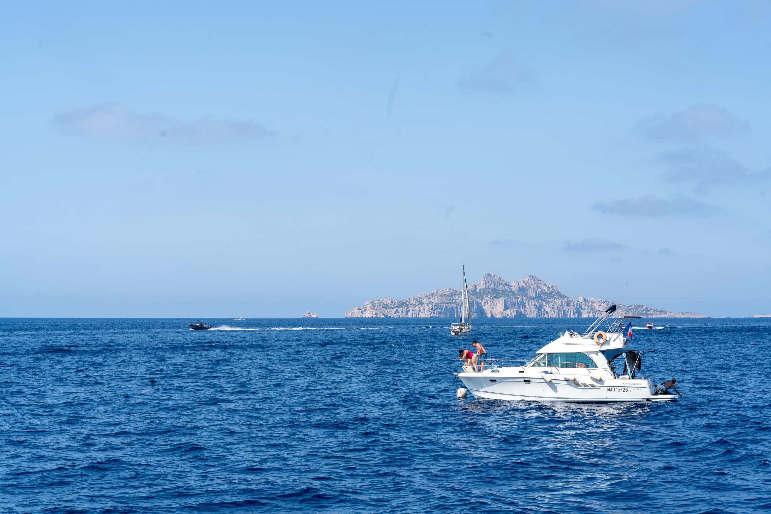 On the open sea under the blue sky, a white yacht with a cabin is sailing in the front right of the frame. Two people are standing on the stern of the boat, and there is a retracted mast on the deck. In the middle of the frame is an island with exposed rock walls and a flat top. There is a sailboat and several small boats in front of the island. The sea is dark blue with fine waves, and there are a few thin clouds in the sky.