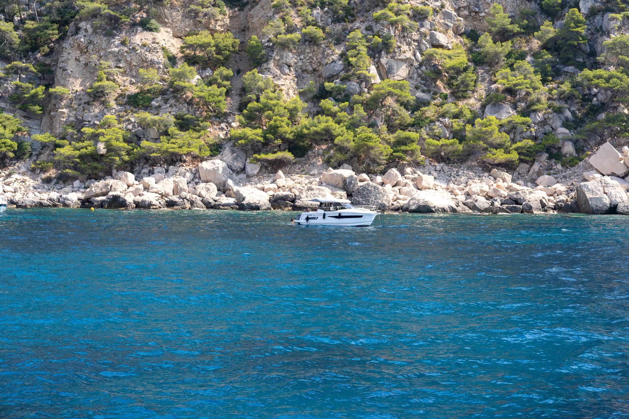 A section of steep coast: the upper half of the frame is a light gray rock wall, with dense dark green pine trees growing in the gaps in the rock wall. Below the cliff is a breakwater-like revetment made of large white stones; the sea surface is bright blue-green, with fine ripples on the water, and a white yacht is driving slowly along the stone shore.