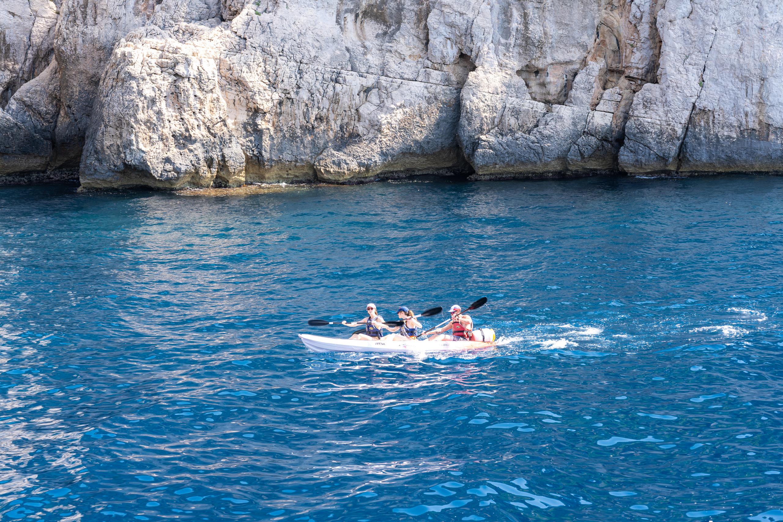 The turquoise water clings to a tall gray-white limestone cliff, which is covered with cracks and hollows. On the water, two people each paddled side by side in a white kayak. One of the three people, wearing red, was waving his oar forward, and the oar stirred up white water spray.