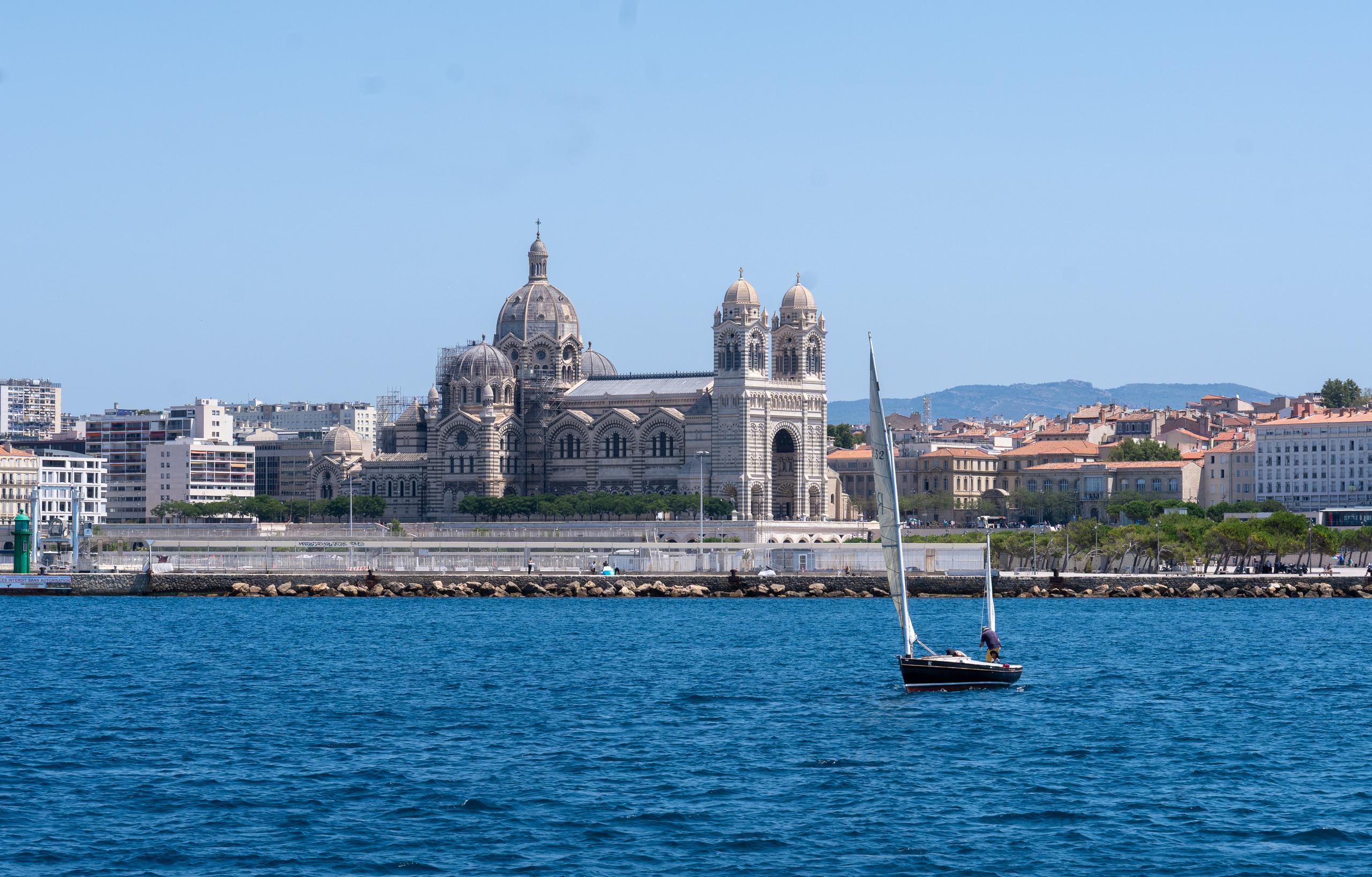 Looking at the city's waterfront from the sea: To the left of the center is a majestic Romanesque church with a huge dome and two square towers. The exterior wall is made of gray-white and dark-striped stone. On both sides of the church are dense urban buildings and red tile roofs. A stone breakwater runs across the front of the church. On the lower right, a white sailboat is sailing across the dark blue sea.