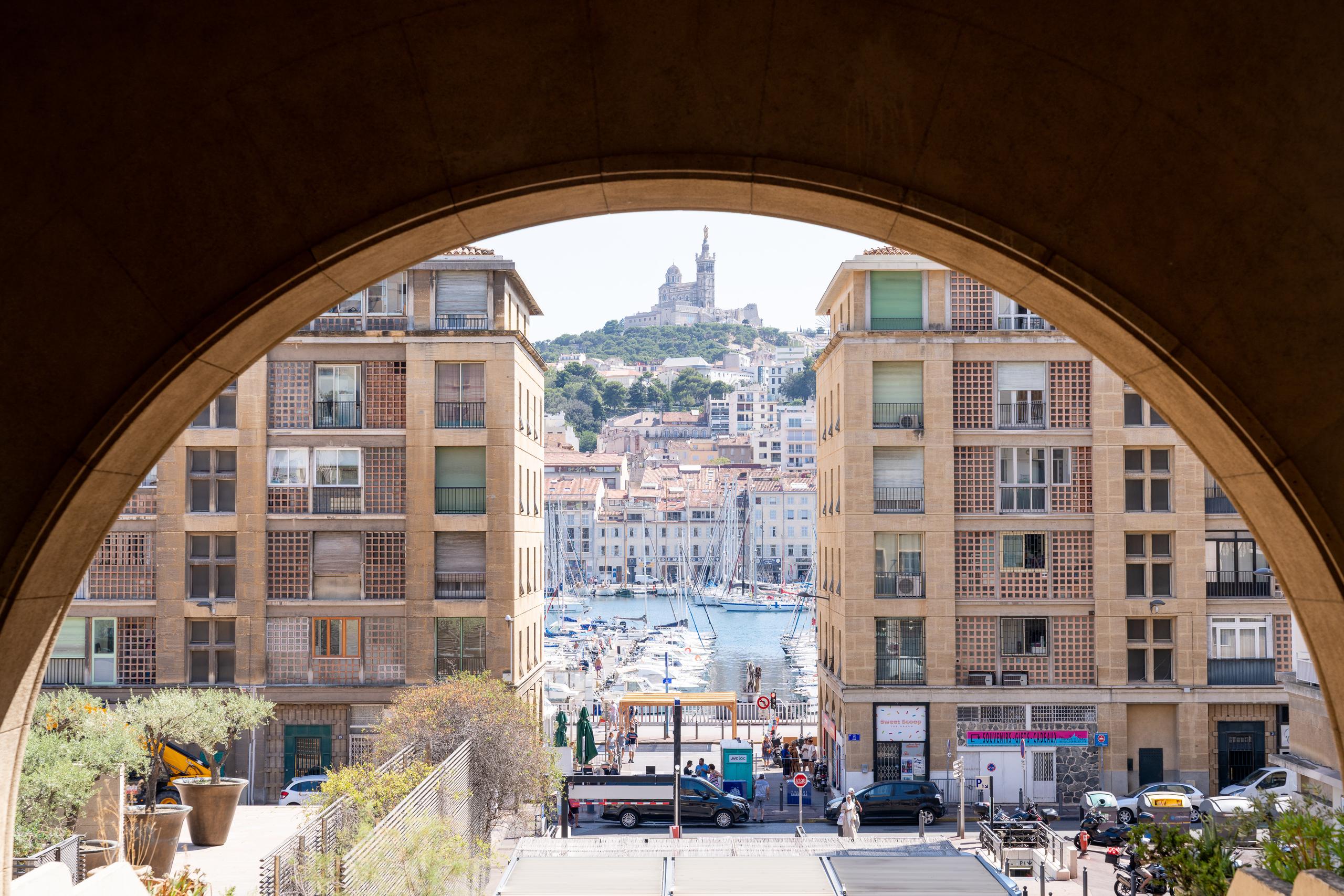 Looking out from inside a huge stone arch: outside the arch, there are modern apartment buildings with beige square veneers on both sides. Through the middle of the arch is a street leading down to the harbor. The harbor is full of sailboats, and there are pedestrians, cars and parked vehicles on the street. On the hill at the end of the street stands a large church-like building with a tall tower and dome. Houses are scattered on the mountainside, and there is mist in the sky.