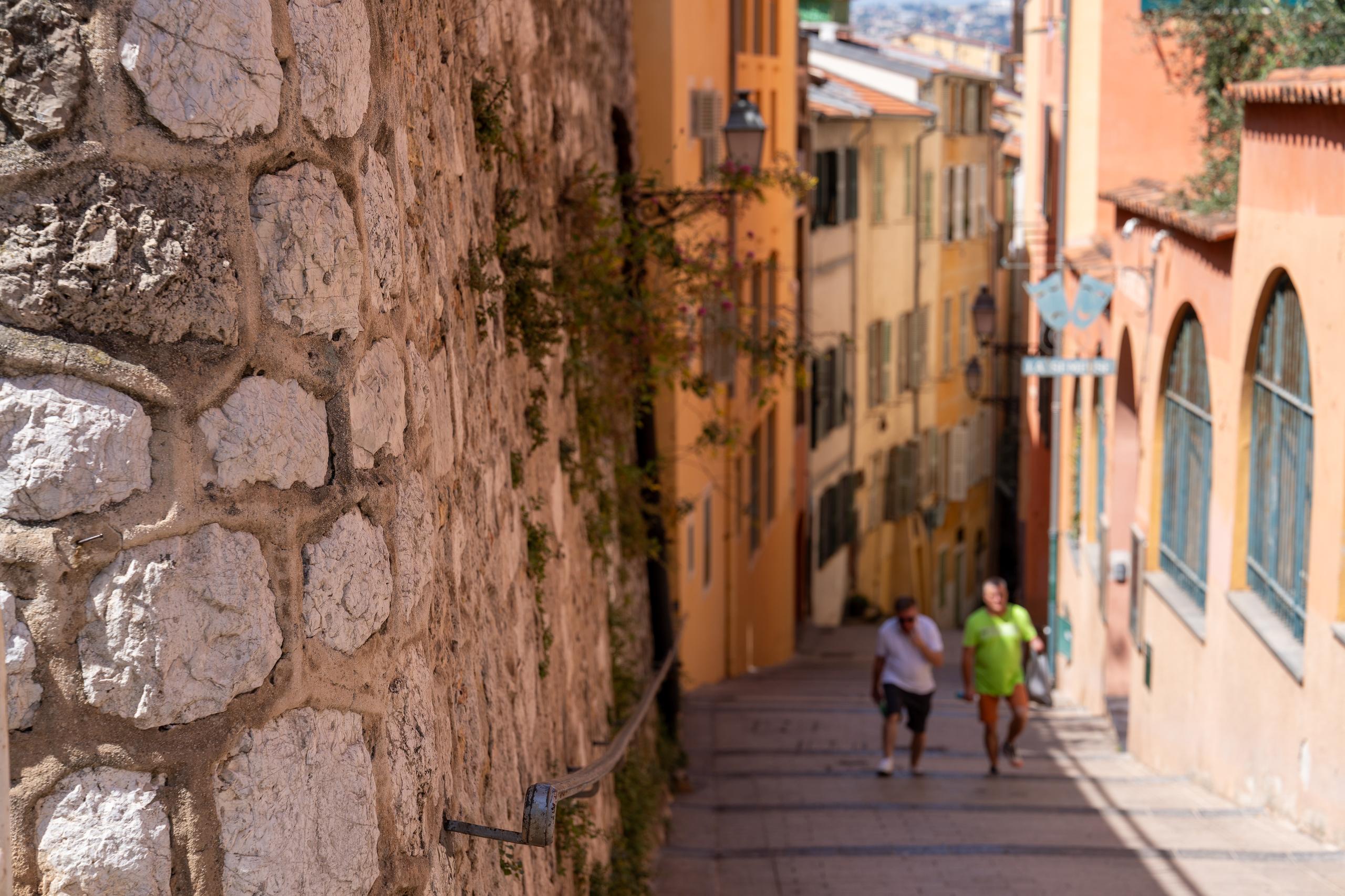 There is a narrow and steep ramp in the old city. On the left side, an entire old stone wall made of irregular gray and white stones takes up almost half of the frame. On the right side are houses with yellow-orange exterior walls. There are shutters and blue window frames on the walls. Two men are walking up the ramp side by side. The ground is a worn stone road, and the background is blurred.