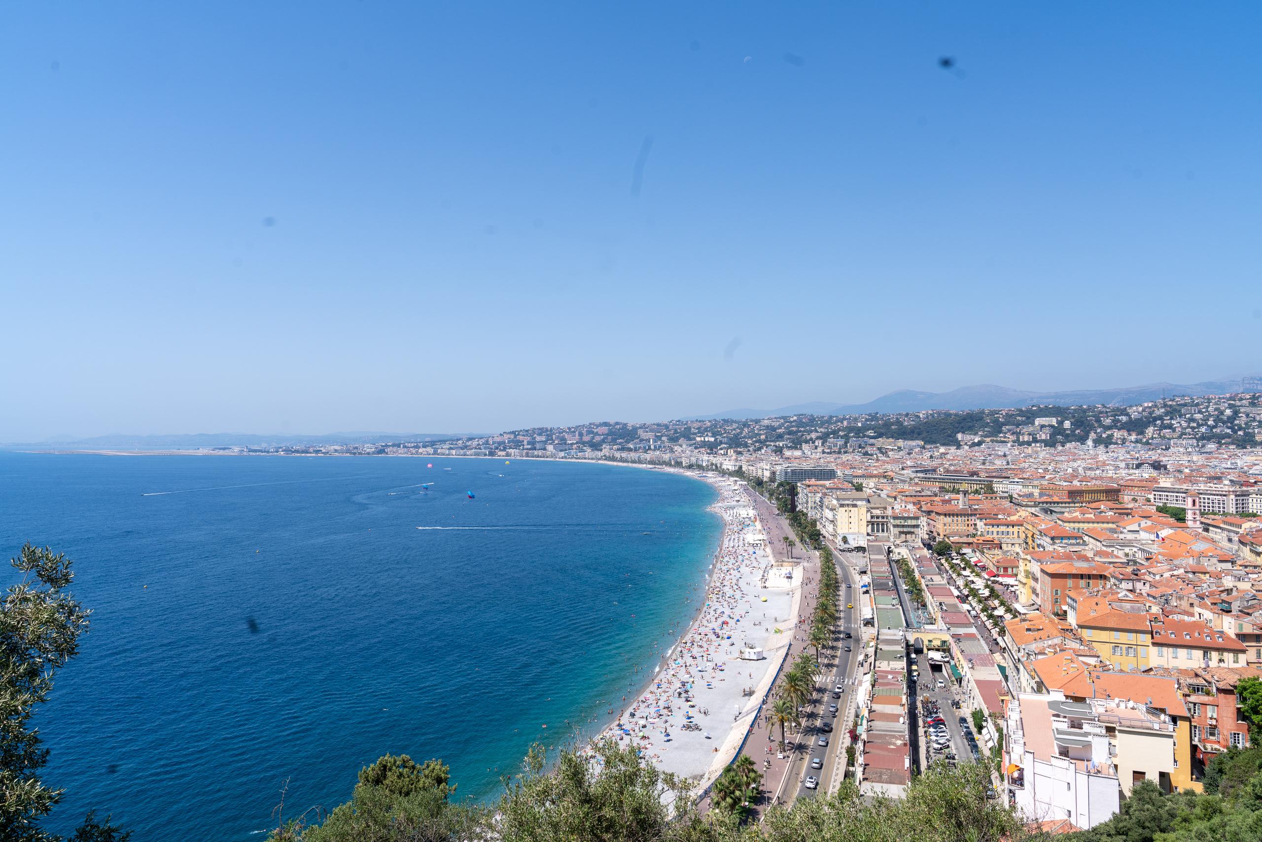 A panoramic view of the bay city from a high angle: a huge arc-shaped beach curves gracefully along the coast, and the beach is densely packed with umbrellas and people; the sea water is bright blue near the shore, gradually turning into dark blue outward; the inner side of the beach is lined with street trees and highways, and further inside is a dense urban area with red tile roofs. There is a green tree crown at the bottom of the frame, and the sky is light blue with mist.