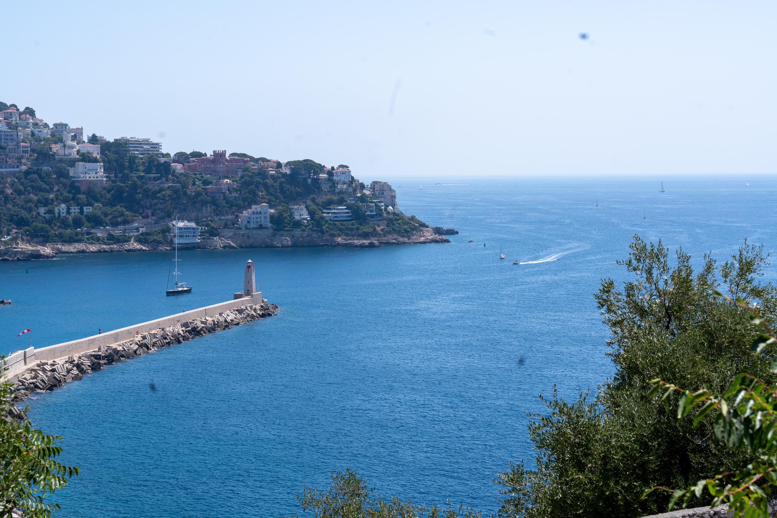 A small bay overlooked from a high place, the sea surface is bright blue; on the left is a stone breakwater extending into the sea, with a small white lighthouse on the head of the breakwater, and several small boats and a sailboat parked inside the breakwater; behind the breakwater is a hillside covered with pine trees and dotted with white and pink houses. On the right are dark green trees in the foreground, and there are several small boats on the sea in the distance.