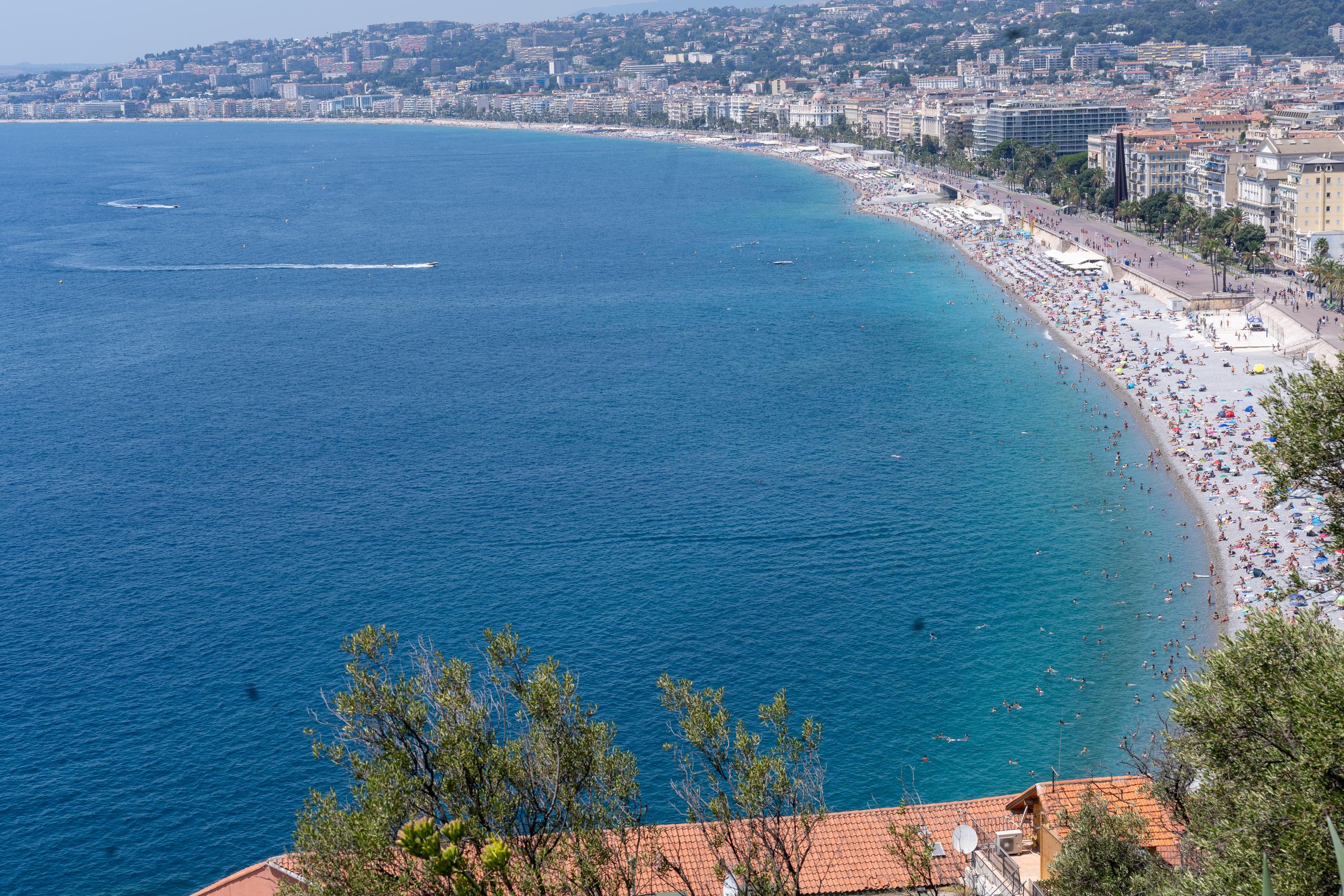 A view of the bay from a high angle: the curved beach extends along the coast, and the beach is crowded with parasols, tents and people playing in the water; the water gradually changes from the emerald green on the shore to the deep blue of the open sea, with a long white wake dragged by a motorboat and a few swimmers on the sea; behind the beach is a coastal avenue and rows of light-colored buildings with red tile roofs. There is a tree crown and a red tile roof at the bottom of the frame.