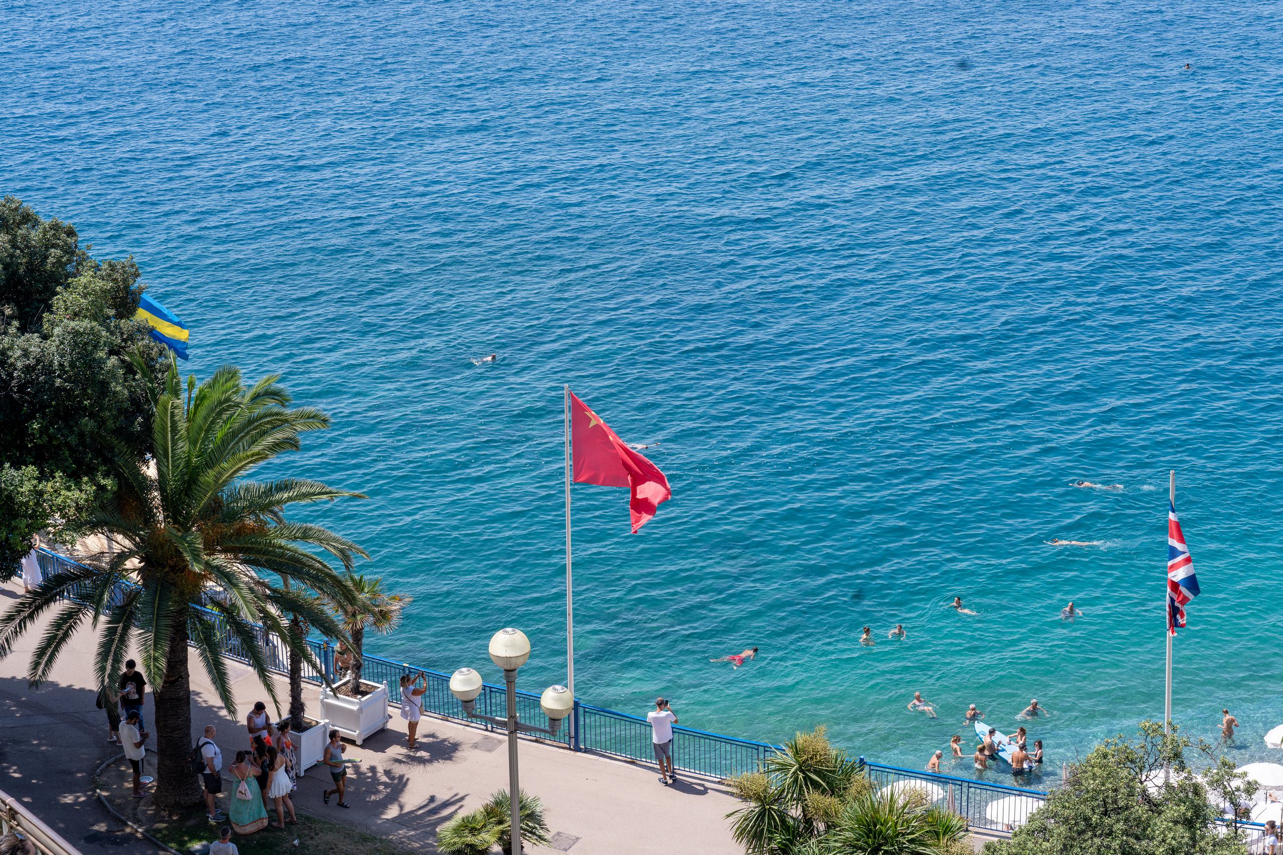 Overlooking the beach: the upper half of the frame is a blue-green sea, with many swimmers scattered in the nearshore water, and inflatable rafts floating on the water; there is a paved walkway on the shore, with several flagpoles standing, flying yellow and blue flags, red flags and British flags respectively. Crowds gather on the walkway and under the shade of the trees, and there are palm trees and street lights on the left.