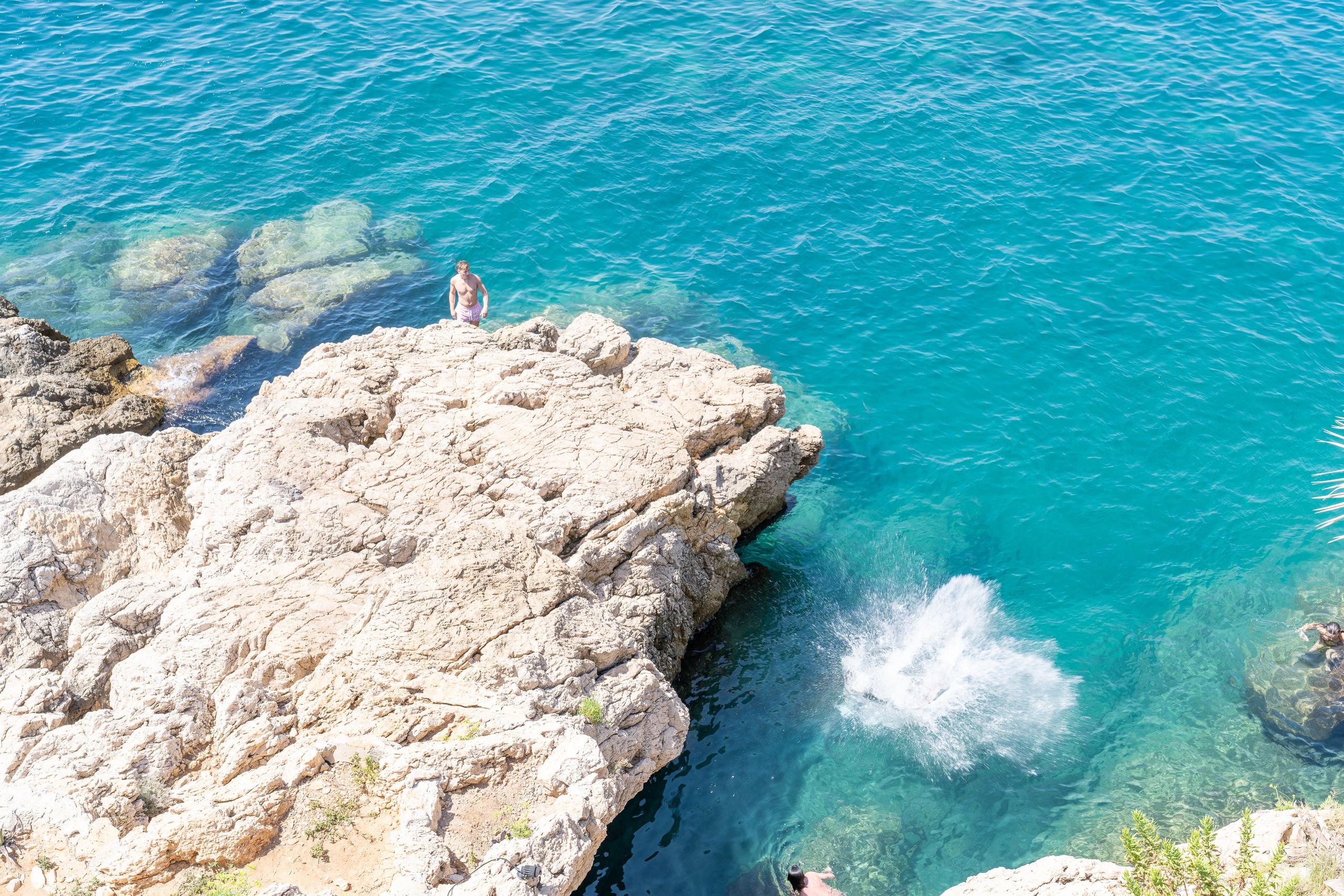 A high-angle overhead shot of a rocky bay: on the left side of the frame is a large flat reef that has been tanned to a light yellow color. A man wearing only swimming trunks is standing on the edge of the rock, facing the water on the right side of the frame; on the right side of the reef is clear turquoise water, and underwater rocks can be seen from the surface of the water. There is a freshly splashed white water in the water; there is another swimmer in the lower right corner of the frame.