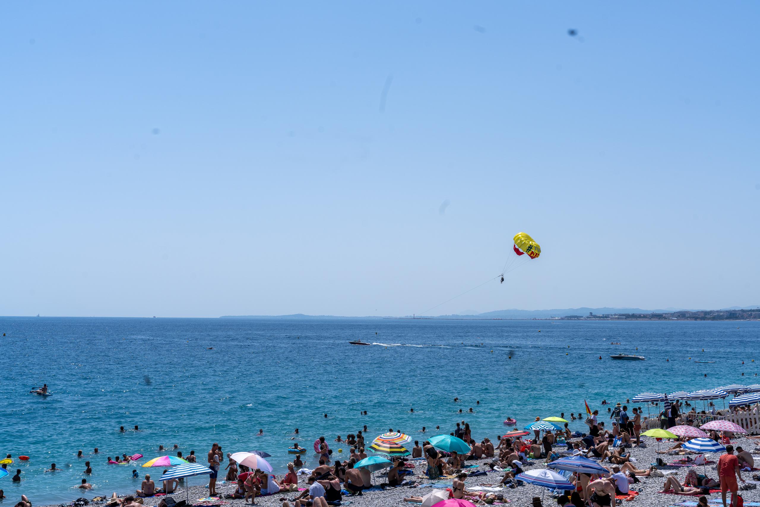 Lively bathing beach: The lower half of the frame is a pebbly beach, crowded with colorful parasols and people lying and walking. There are also a large number of people swimming in the sea; in the sky above the sea to the right, a tourist is being towed by a speedboat for a sea parachute flight, with a person hanging under a colorful parachute; in the distance, the sea level is flat, and the sky is clean and light blue.