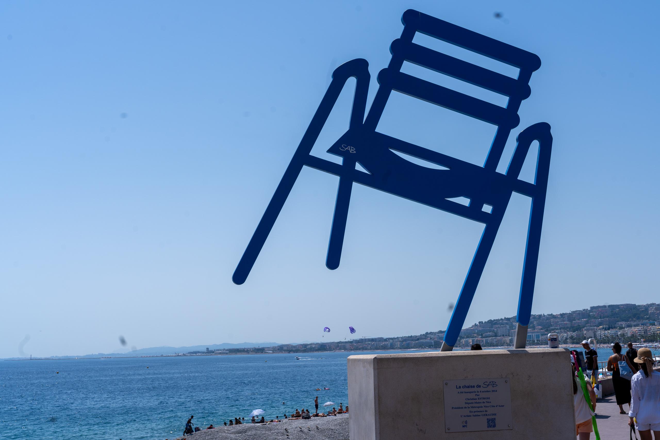In the center of the frame is a giant dark blue sculpture. It is shaped like a slatted chair with the legs turned up and the seat facing the side. It is installed on a square concrete base. There is an explanation board on the front of the base. The sculpture overlaps with the city coastline, sea and beach in the distance. On the lower right are pedestrians on the seaside promenade.