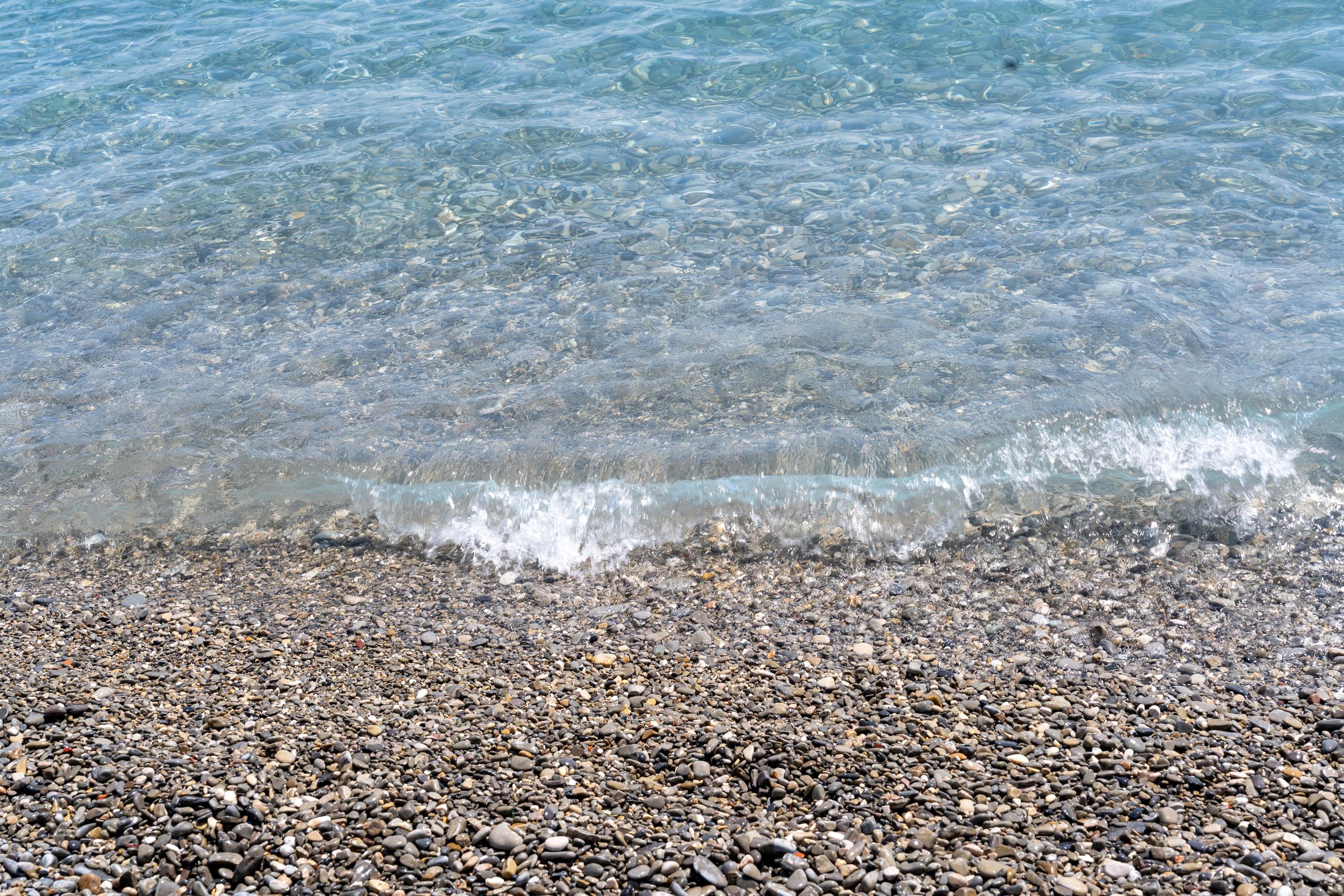 A close-up view of the junction of the water surface and the beach: the upper half of the frame is crystal clear sea water, and the underwater pebbles can be seen through the water surface, and a small white wave is hitting the shore; the lower half of the frame is a gravel beach densely covered with gray-brown pebbles. The pebbles are of different sizes and are washed very smooth by the water.