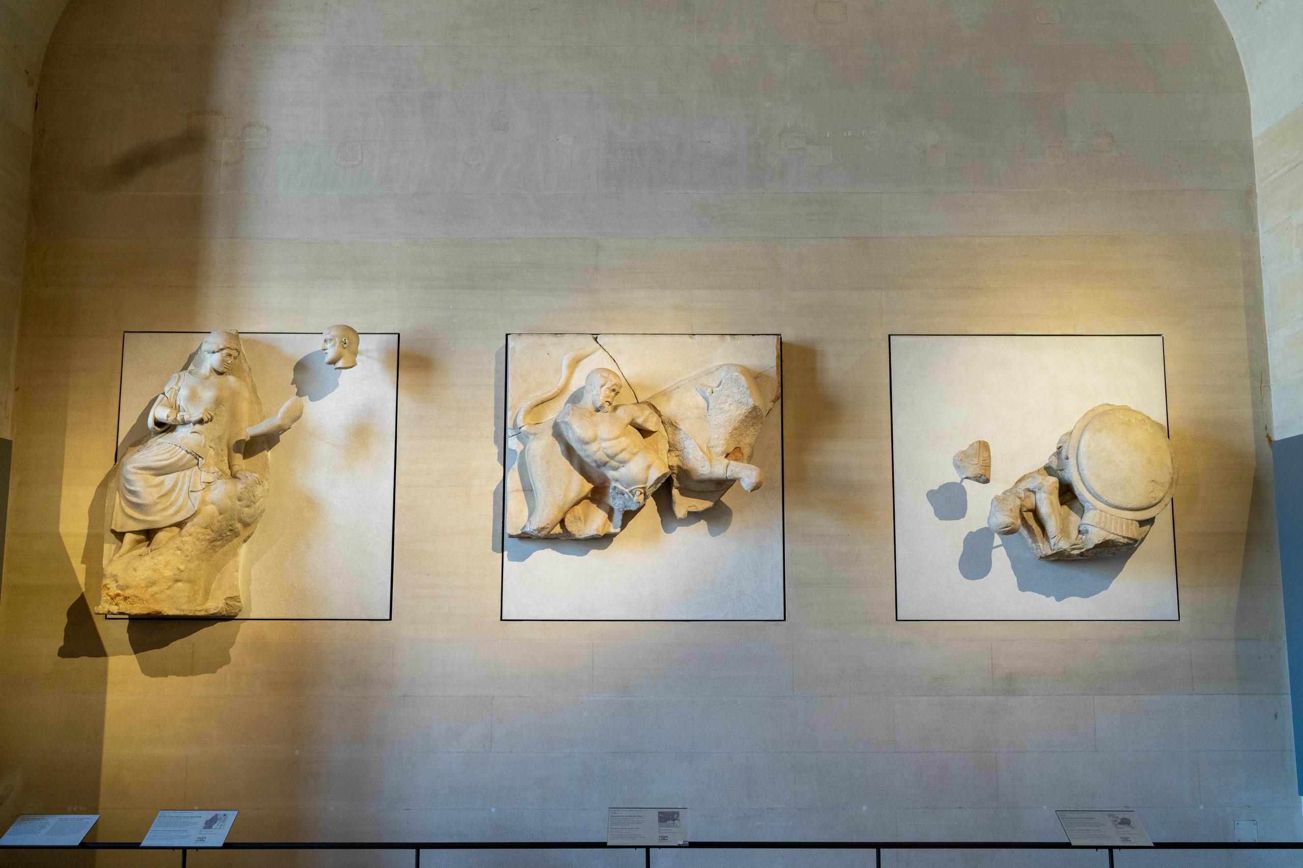On a light-colored stone wall in the museum exhibition hall, three square ancient stone carving fragments are hung side by side. They are off-white and obviously weathered: the one on the left is a relief of a seated figure, and a fragment of a small human head hangs alone on the upper right; the one in the middle is a relief of a half-lying figure; the one on the right is a figure lying on his back and a fragment of a round shield; each exhibit has a small explanation board attached below it.
