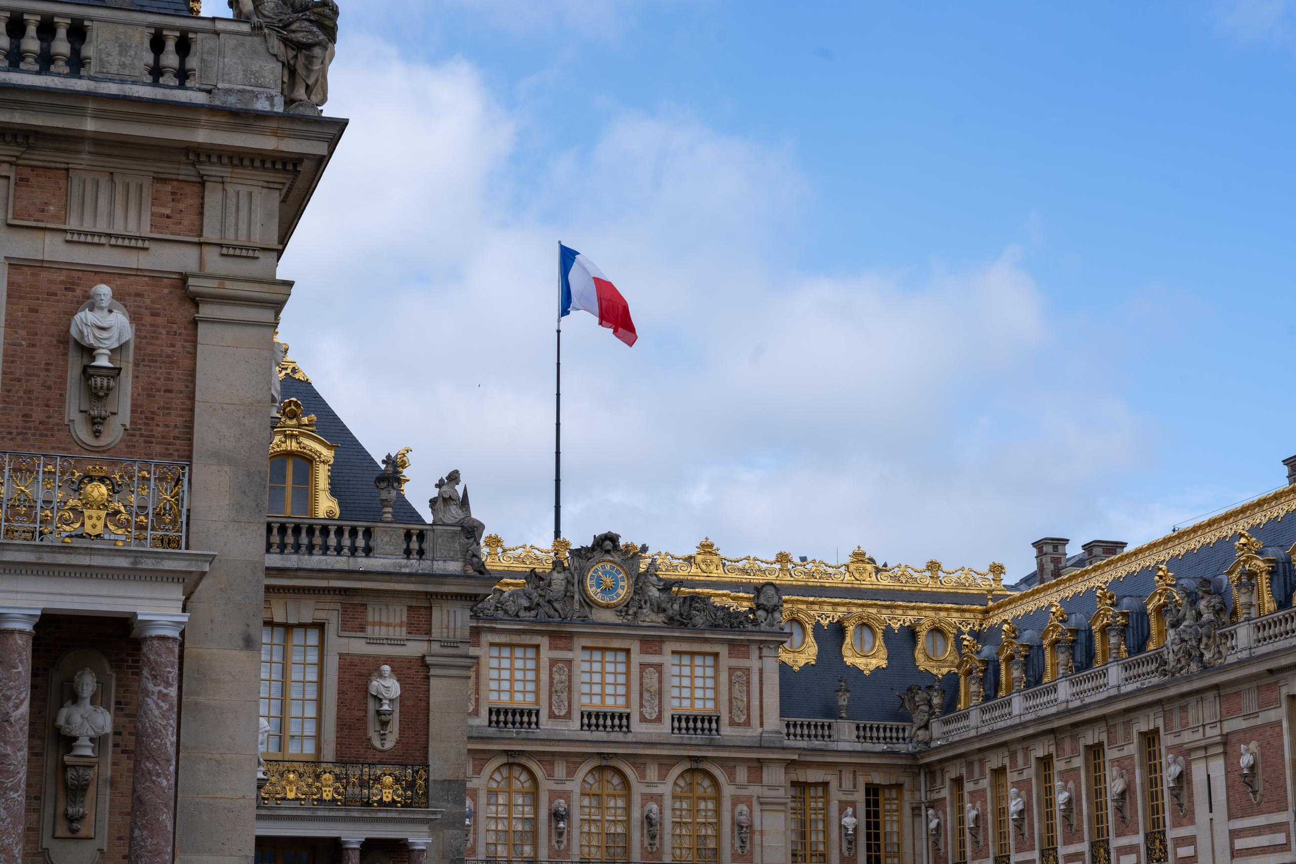 Under the blue sky and white clouds, a corner of a magnificent palace building: the outer walls are made of alternating red bricks and light-colored stones, the roof is made of dark slate, and the cornices, window frames, railings and roof edges are decorated with a large number of gilded decorations; a blue, white and red tricolor flag floats on a flagpole high in the center; a white human bust is placed in the niche of the tower on the left, and there are stone statues at the corners of the railings.