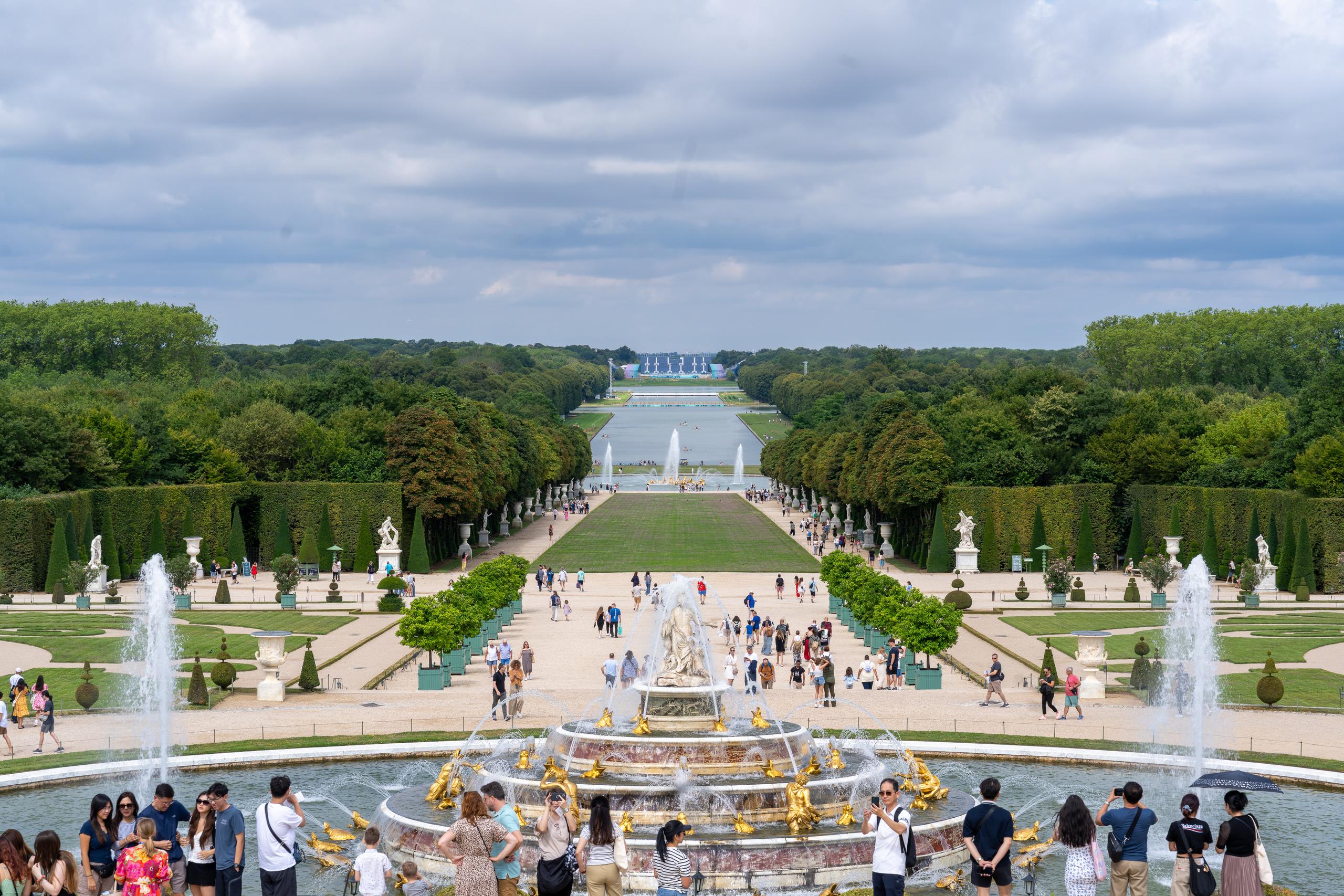 A regular French garden is overlooked from a high platform: directly in front is a central axis, flanked by geometrically trimmed lawns, hedges and rows of potted trees; at the end of the axis is a large pool, with many tourists scattered on the grass and walking trails; in the foreground is a circular fountain decorated with gold-plated sculptures, and the water jets form high water curtains on both sides of the frame.