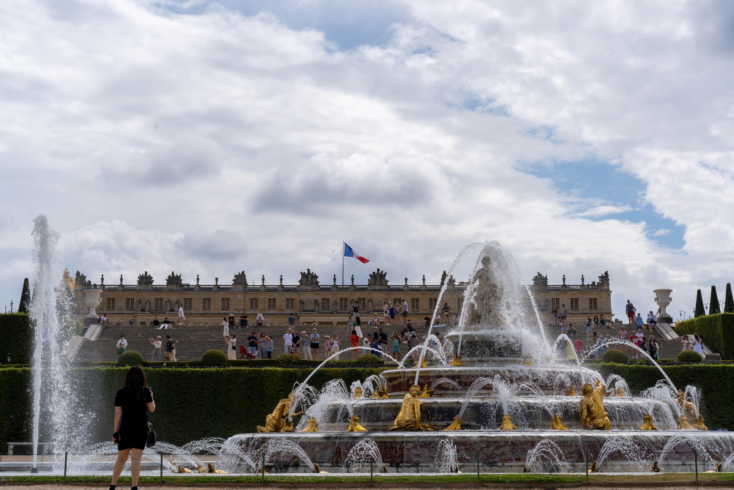 Fountain in the palace garden under a cloudy sky: on the lower right is a large multi-layered fountain. There are many gilded figures and animal sculptures standing on the edge and upper floor of the pool, with multiple arc-shaped water columns shooting out from their mouths; behind the fountain is a wide stone staircase and the palace facade. A tricolor flag flies in front of the palace, and many tourists gather on the stone steps; on the left side there is a high spray of water column.