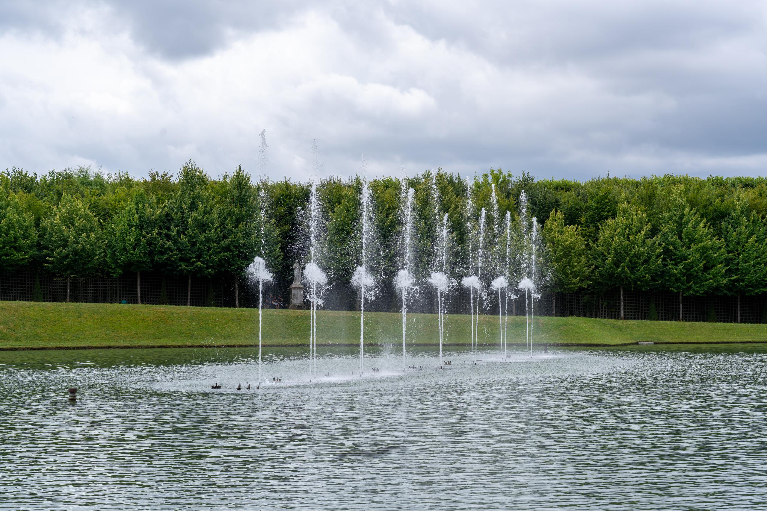There is a row of nozzles spraying water in a large pool. About ten thin and tall water columns spray neatly into the air and fall back to the water surface to form white water spray and mist. Behind the pool is a gently sloping lawn. Above the lawn are tall green trees neatly trimmed and arranged in rows, forming a green wall. There are stone guardrails in front of the trees. The sky is filled with thick gray clouds, and there are several water birds on the water.
