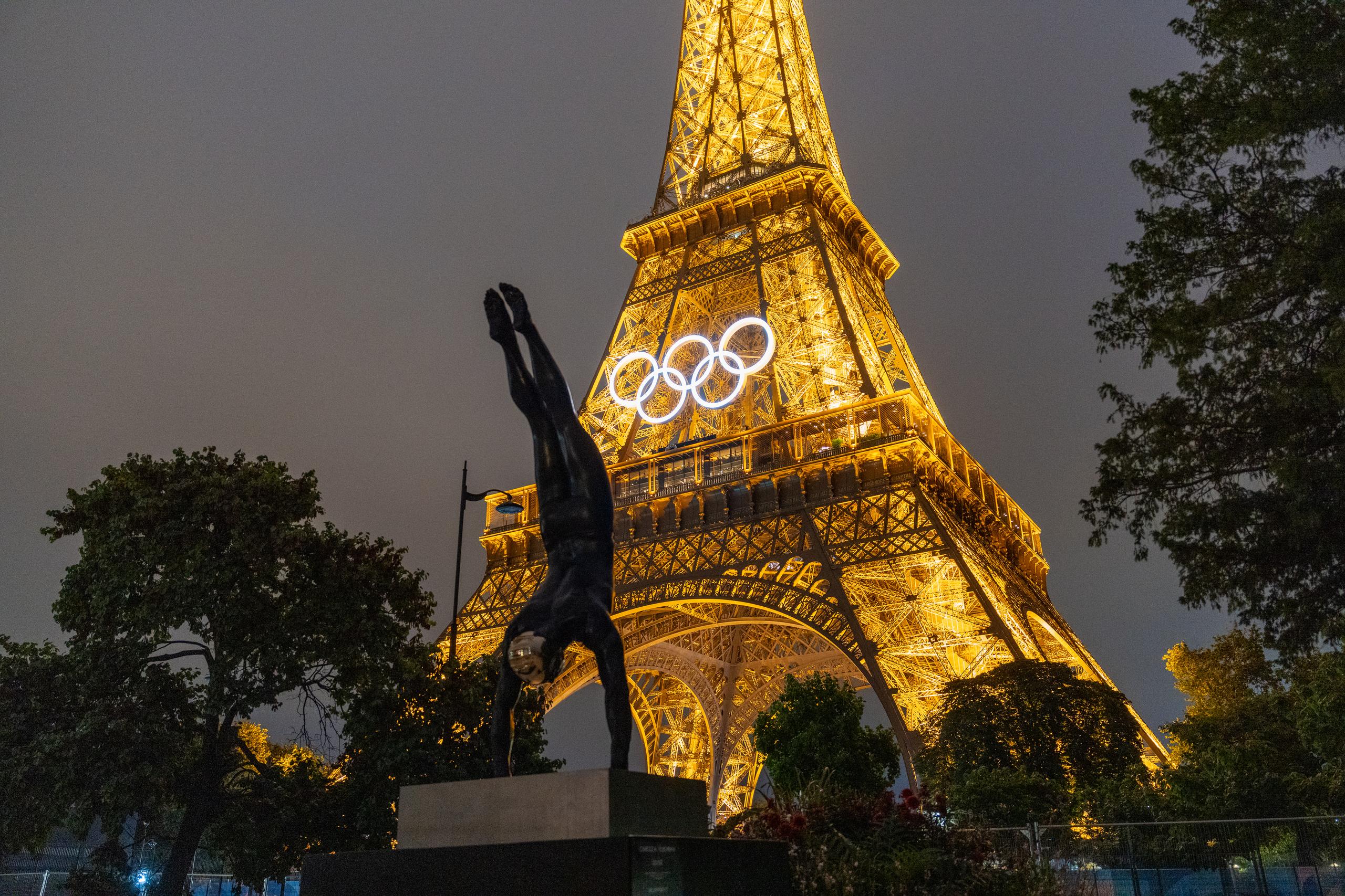 There is an iron tower in the night scene. The entire tower is illuminated by golden lights, and a white five-ring pattern composed of luminous lines is hung on the tower; in the foreground on the left side of the frame is a black gymnastics figure statue, an inverted human sculpture standing on a square base, surrounded by silhouettes of dark trees, and there are branches on the right side of the frame; the sky is a thick gray-blue, and the overall lighting atmosphere is at night.