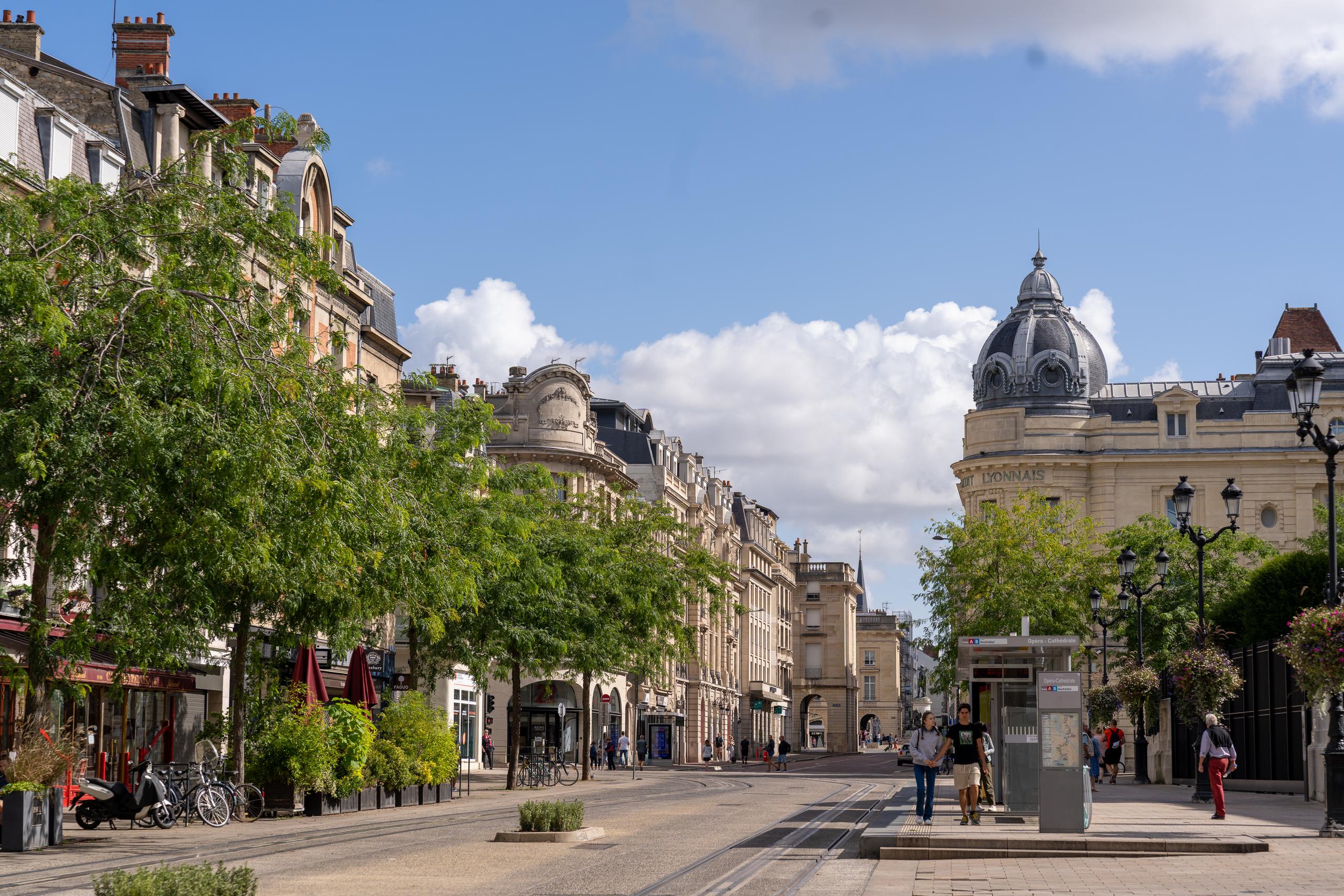 European city street on a sunny day: On both sides are multi-story classical buildings made of beige stone and brick, with balconies and painted window decorations on the walls. On the left side of the building are shops with red awnings on the first floor; on the right side is a very eye-catching corner building with a dark gray dome and tower; in the center of the street is paved with light-colored stone slabs and embedded with tracks, and there are several pedestrians on the road; on the left side there are rows of large pruned trees, and the sky is blue.