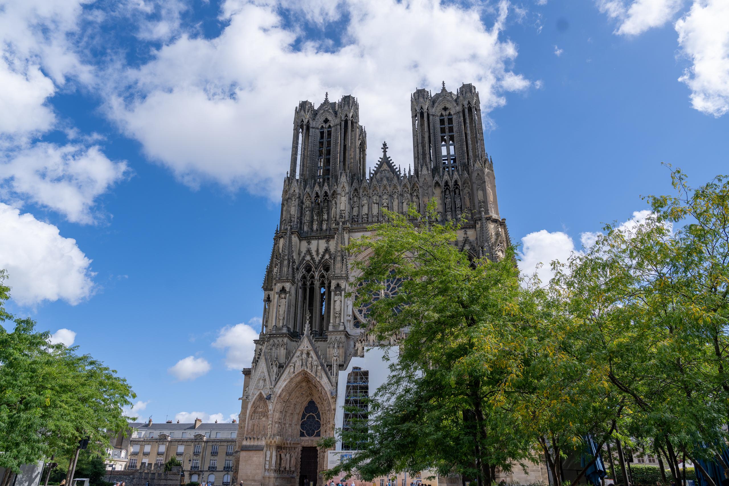 Looking up at the front of a magnificent Gothic cathedral: the church has two symmetrical towers, three pointed arch openings, a rose window in the center, dense stone carving details, and the overall color is grey-brown; the right side of the church is blocked by scaffolding and white enclosures and is under repair; the foreground and right side are lush green trees, partially blocking the lower part of the church, and the sky is blue and covered with large white clouds.