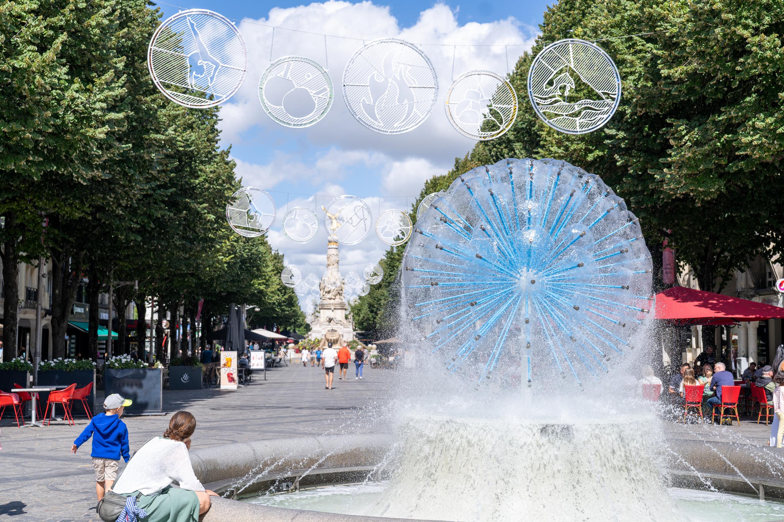 City Pedestrian Square during the day: To the right of the center is a large fountain. The jetted water forms an umbrella-shaped white water curtain, with a circle of radiating blue tubular components in the water curtain. Behind it is a wide pedestrian street, lined with rows of tall green trees and shops, with strings of round white hollow garlands hanging between the trees. On the left are a child in blue and a woman sitting by the fountain with her back to the camera.