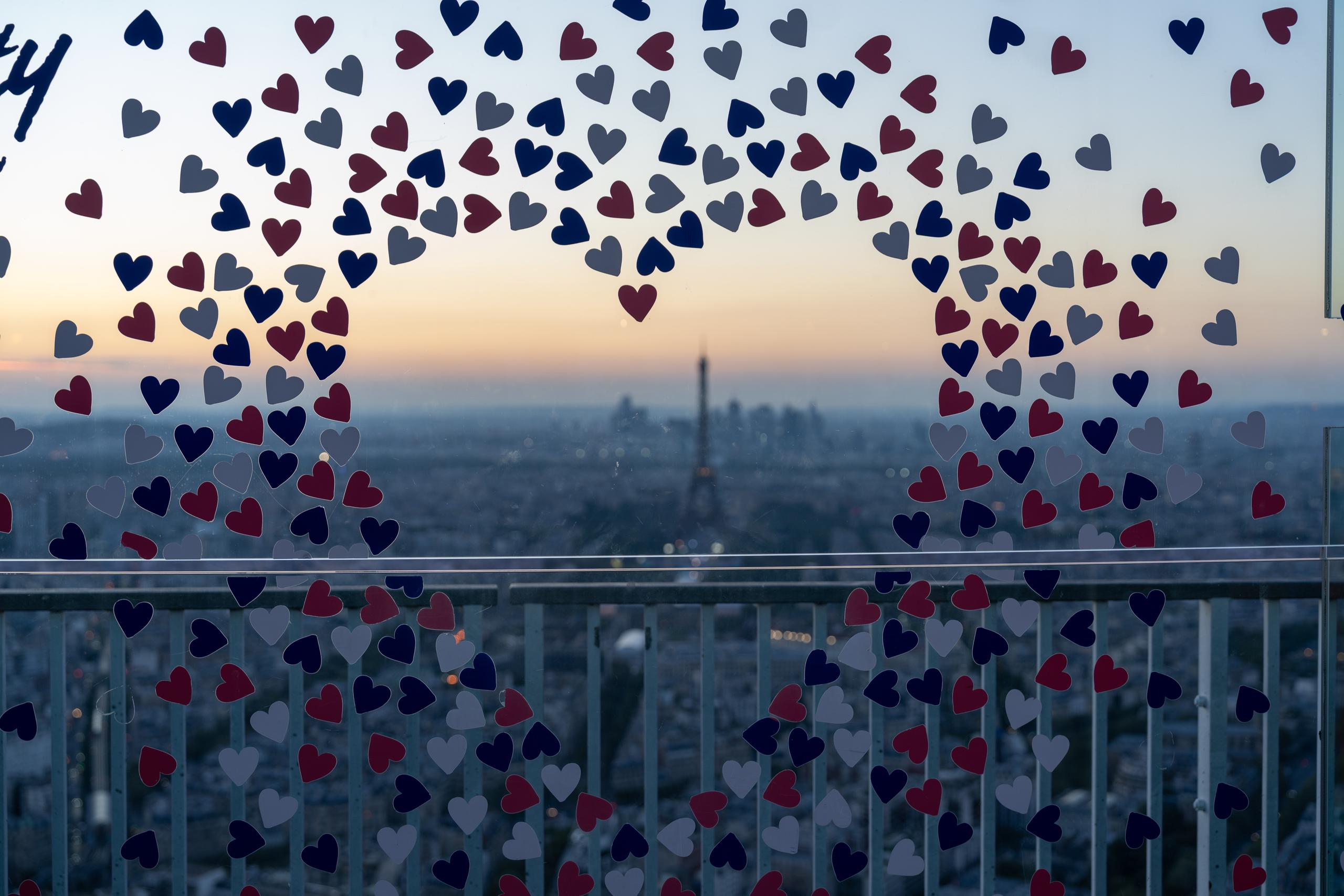 Looking out from behind the high railing: a large number of red, white and blue heart-shaped pieces of paper are pasted on the glass or railing, densely arranged in two clusters of arcs; behind the paper is a panoramic view of the city in the twilight, with an orange-yellow sunset on the horizon, and the silhouette of the iron tower and the vague outline of the city can be recognized in the distance; there is a metal crossbar passing through the lower middle part of the frame.