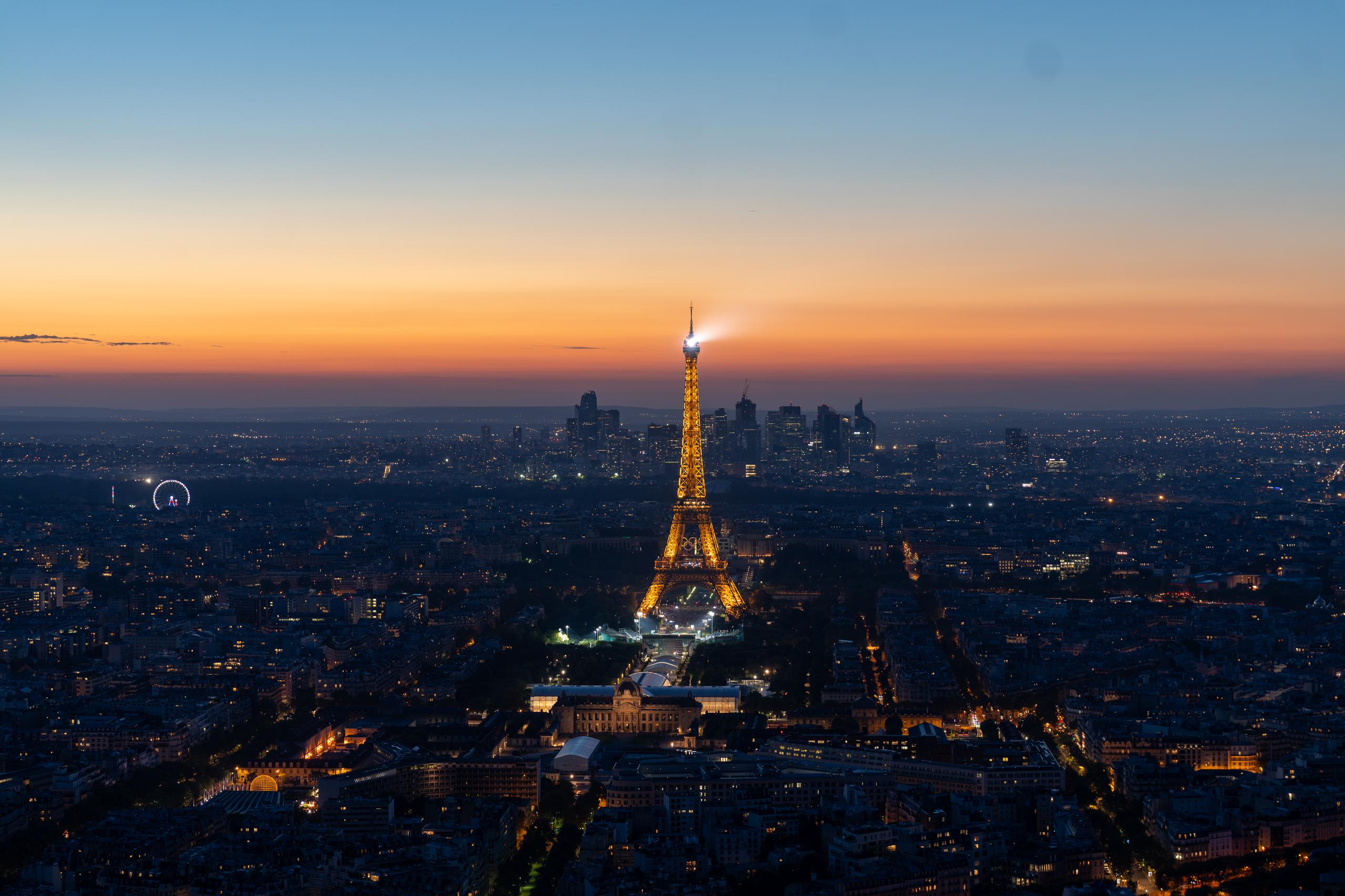 Overlooking the entire city from a height after sunset: the iron tower in the center of the frame is illuminated golden yellow by lights, with a bright flash on the top of the tower; the streets around the tower extend in a radial shape, the city is full of lights, and on the horizon in the distance is a dense outline of high-rise buildings; the sky gradually changes from orange-red at the horizon to dark blue above.